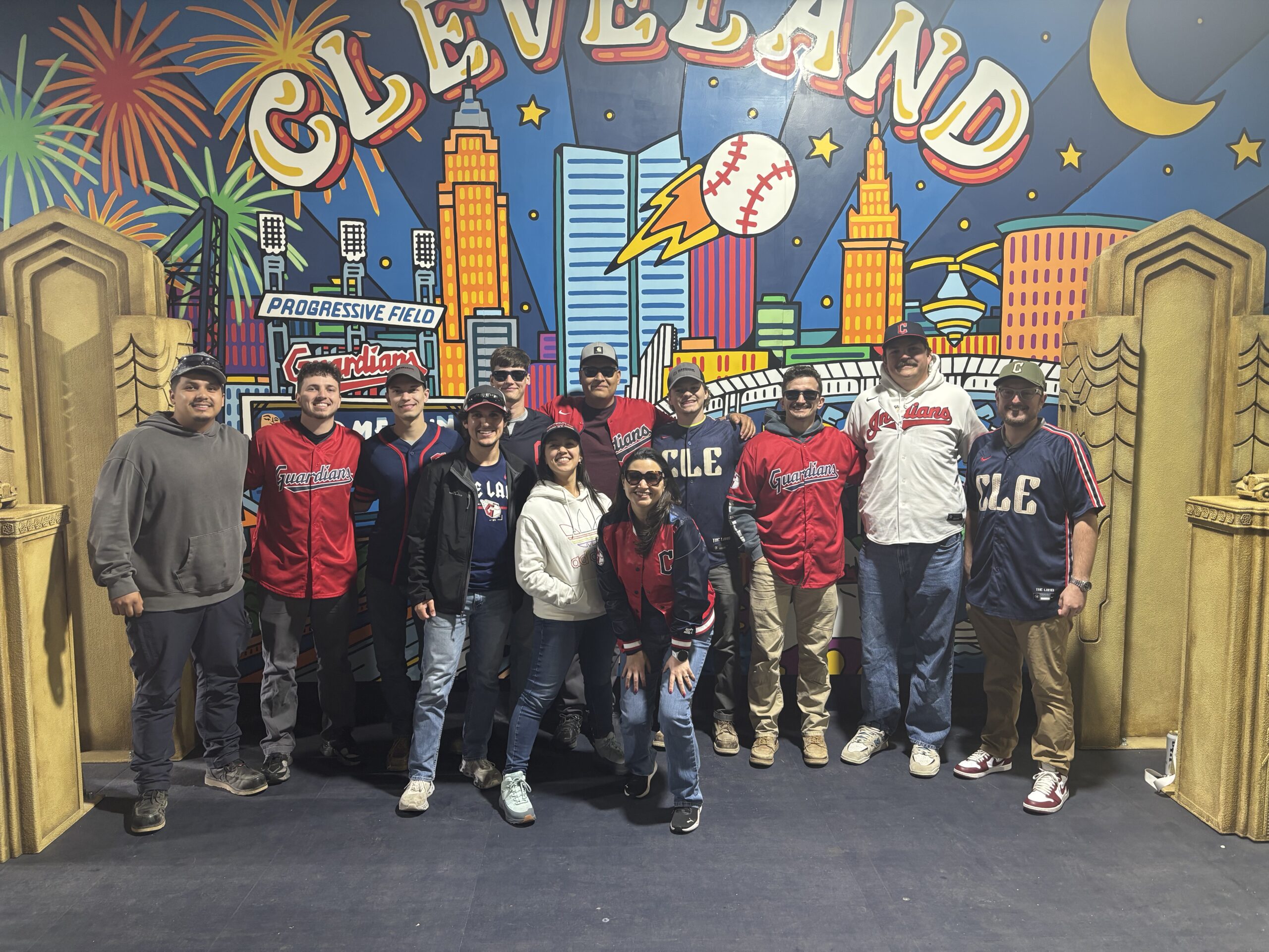 A group of people pose in front of a colorful mural featuring Cleveland, city buildings, fireworks, and baseball imagery. Most are wearing baseball jerseys and caps, smiling at the camera.