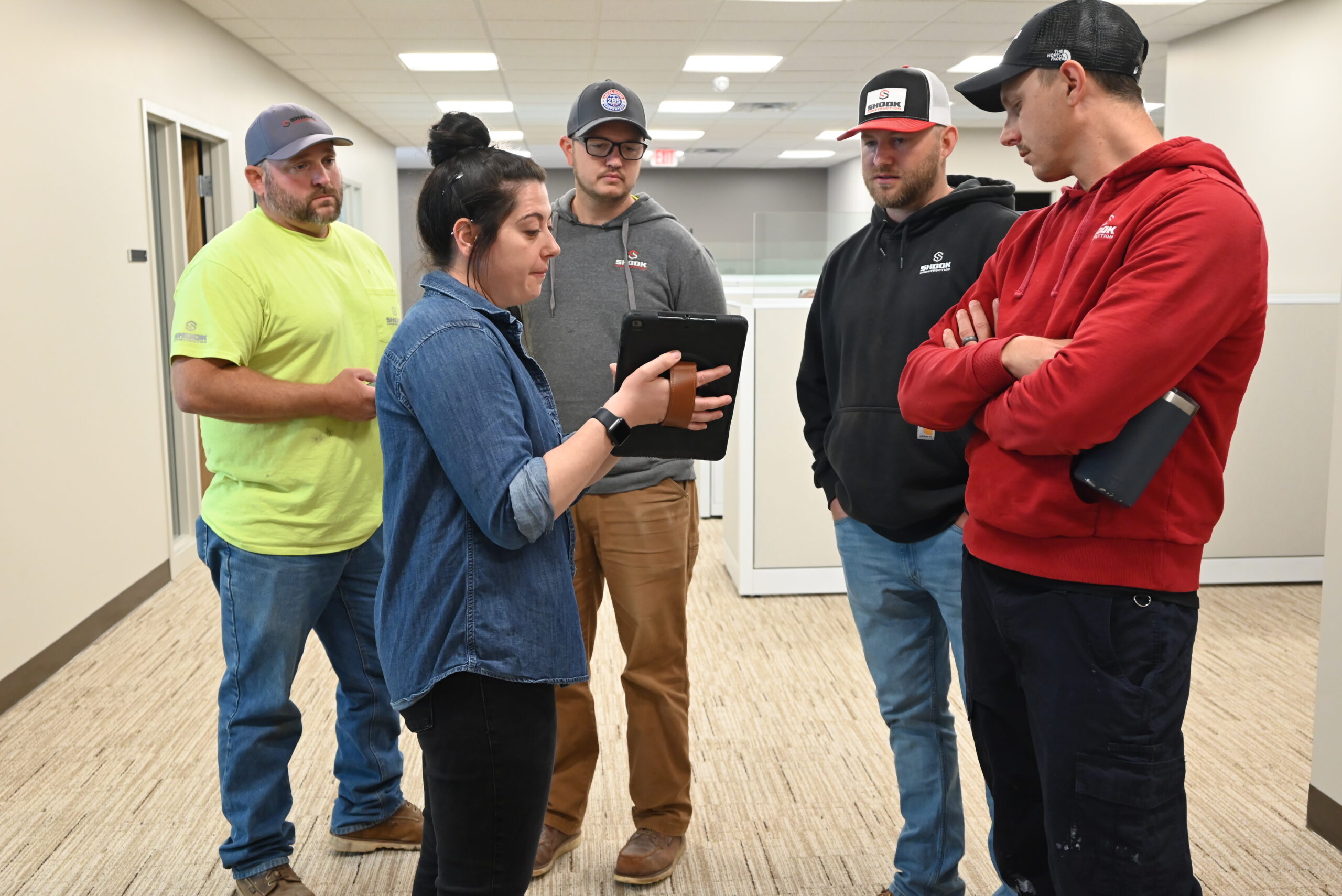 Five people stand in an office hallway. One woman shows something on a tablet to four men, who are listening attentively. Everyone appears to be engaged in discussion.