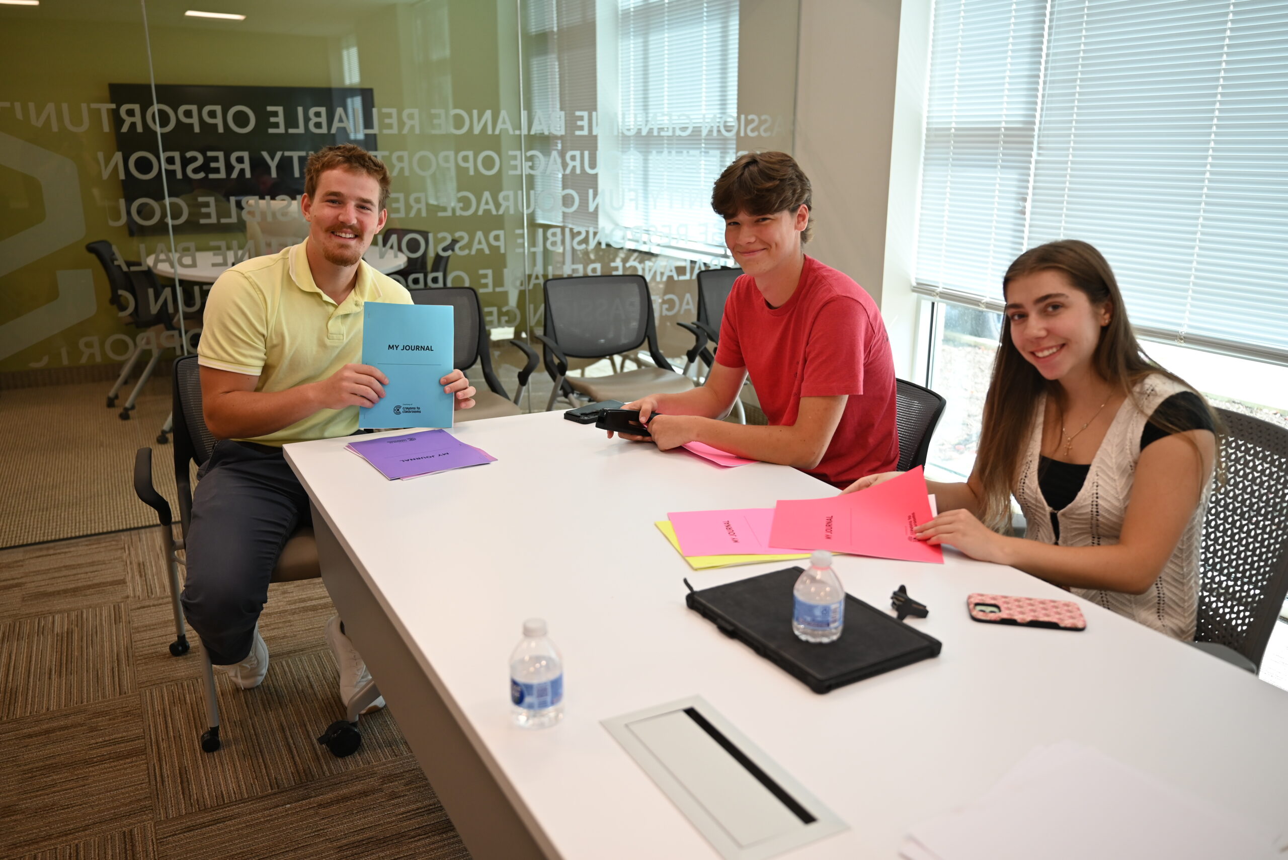 Three young adults sit at a conference table with papers, folders, and water bottles in front of them. They are smiling and appear to be in a modern office or meeting room with large windows.