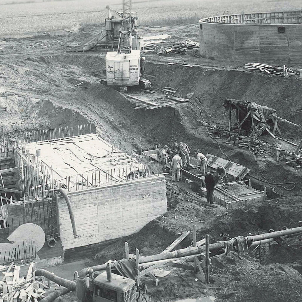 Black and white image of a construction site with workers building concrete structures, rebar exposed, pipes running along the ground, and a large circular tank in the background. Machinery and tools are scattered around.
