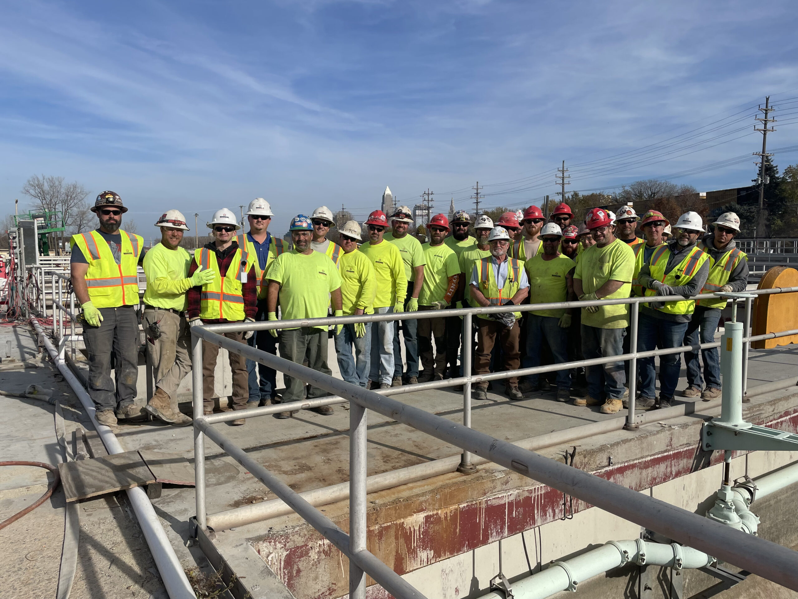 A group of construction workers in safety gear and bright yellow shirts stand together on a work site outdoors, posing for a group photo on a sunny day.