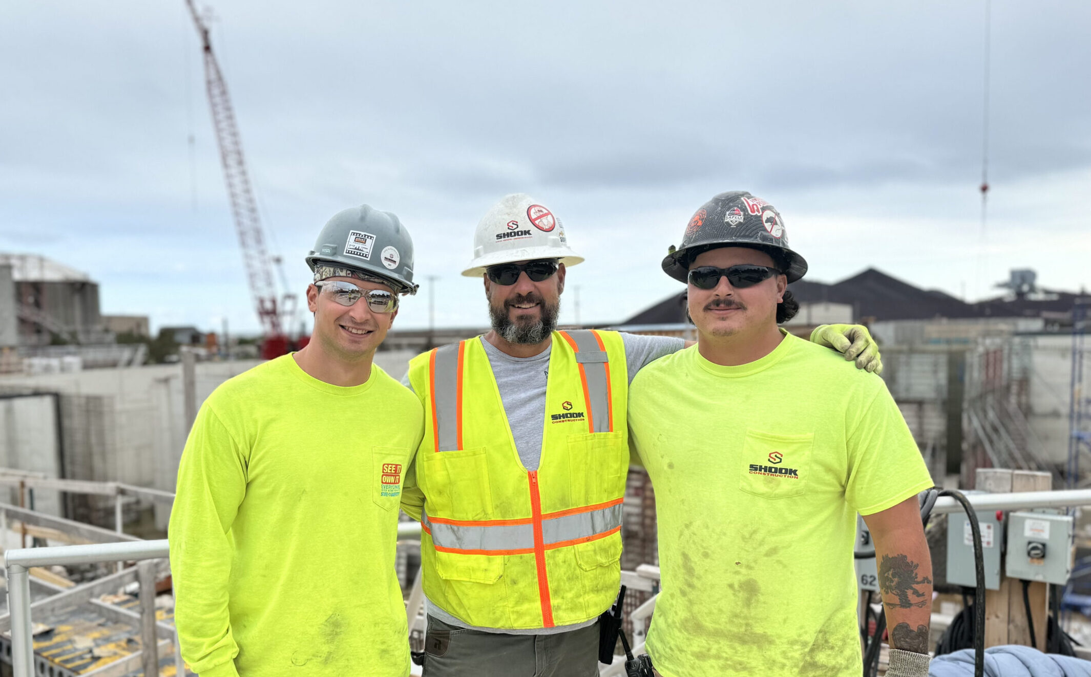 Three construction workers wearing safety gear and neon yellow shirts stand together at a construction site, smiling at the camera. A crane and building materials are visible in the background under a cloudy sky.