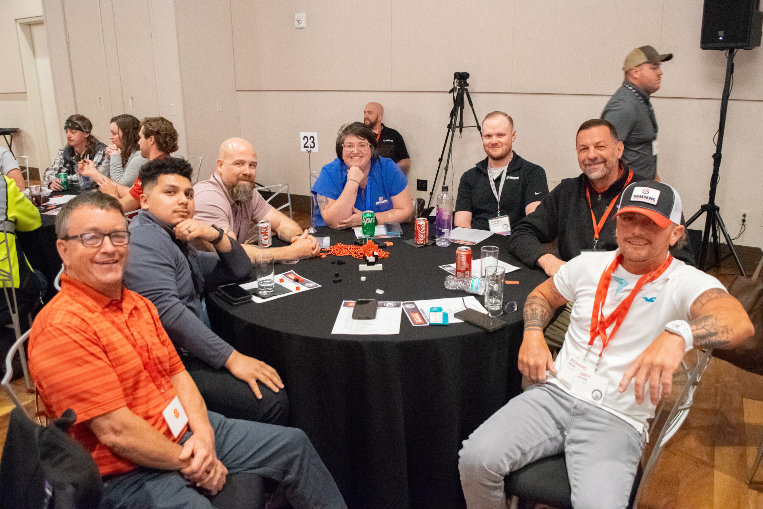 Eight people sit around a round table at an event, smiling at the camera. Papers, drinks, badges, and poker chips are on the table. A camera and tripod are visible in the background.