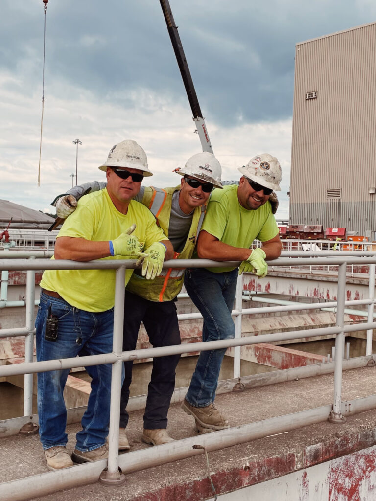Three construction workers in safety gear and neon shirts lean on a metal railing at an industrial site, smiling. A crane and large building are visible in the background under a cloudy sky.
