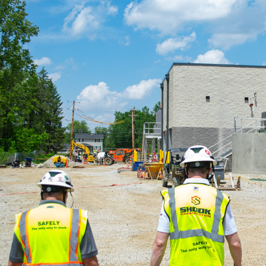 Two construction workers wearing safety vests and helmets walk toward an active job site with equipment, vehicles, and a partially built structure under a blue sky with clouds.