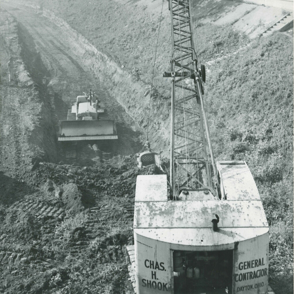 A crane and a bulldozer work in a dirt excavation site. The crane has Chas. H. Shook General Contractor, Dayton, Ohio on its side.