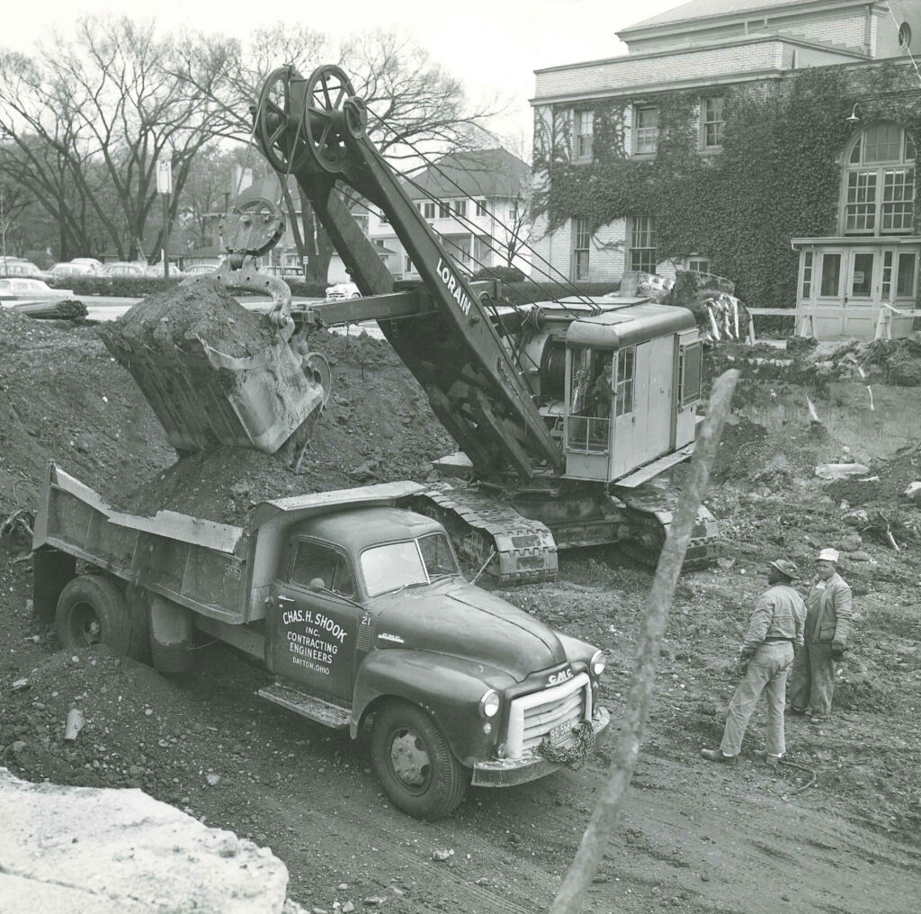 A vintage excavator loads dirt into a dump truck at a construction site while two workers stand nearby. A building covered in ivy and several parked cars are visible in the background.