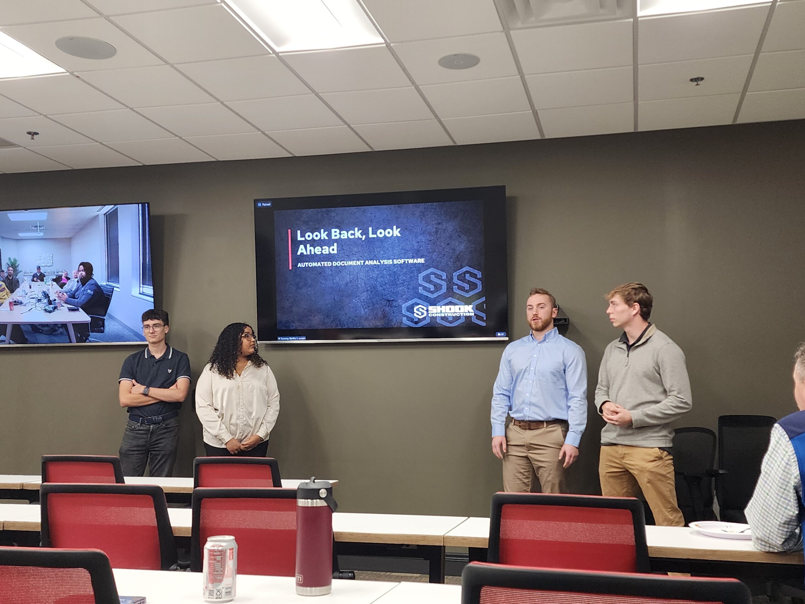 Four people stand in front of a classroom, two presenting near a screen that reads Look Back, Look Ahead. There is another group in a meeting on the TV in the background. Several desks and chairs are visible.