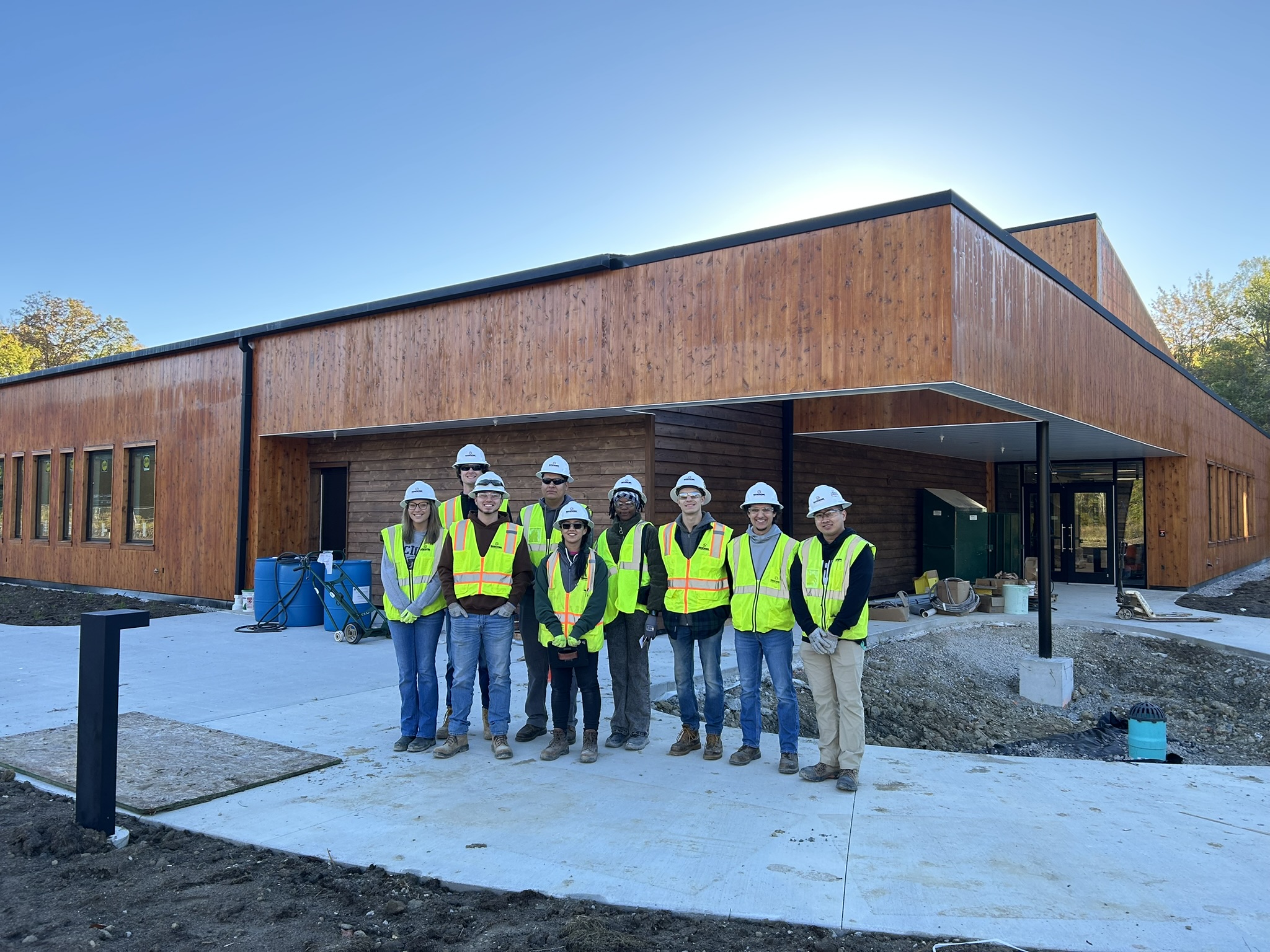 A group of nine people in hard hats and bright safety vests stand in front of a modern wooden building under construction on a clear, sunny day.