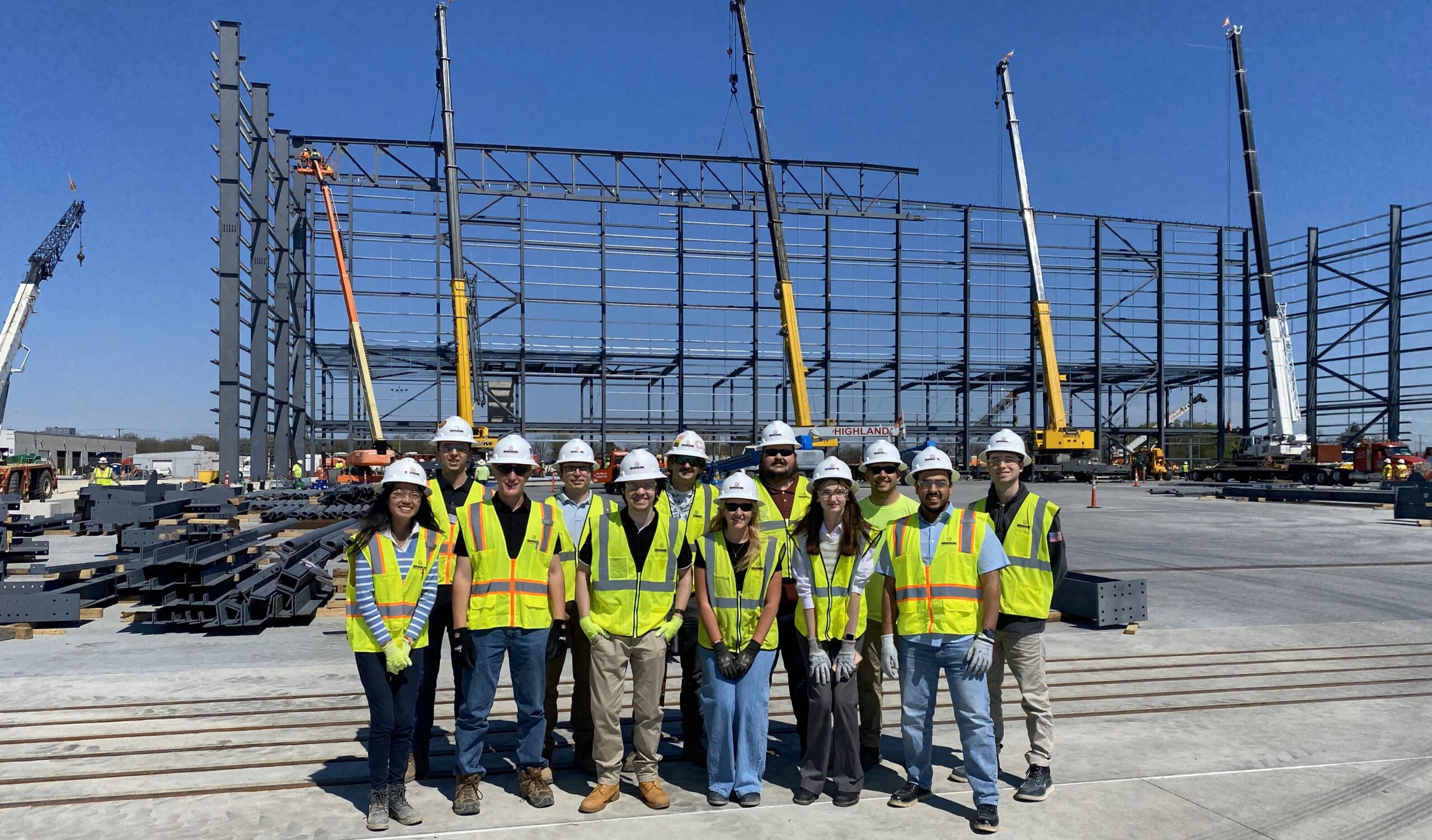 A group of people in safety vests and helmets stand in front of a large, partially constructed steel building framework at a construction site with cranes in the background.