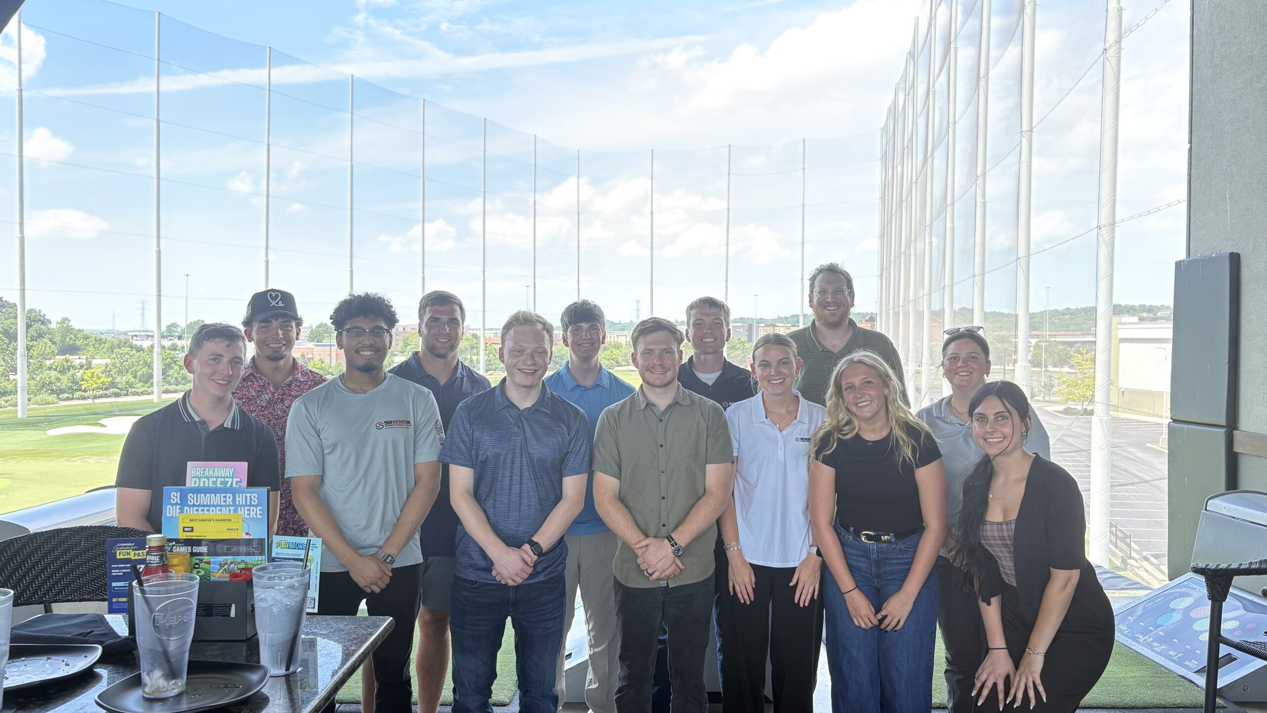 A group of 14 people pose and smile together at an outdoor driving range on a sunny day; nets, green grass, and blue sky appear in the background.