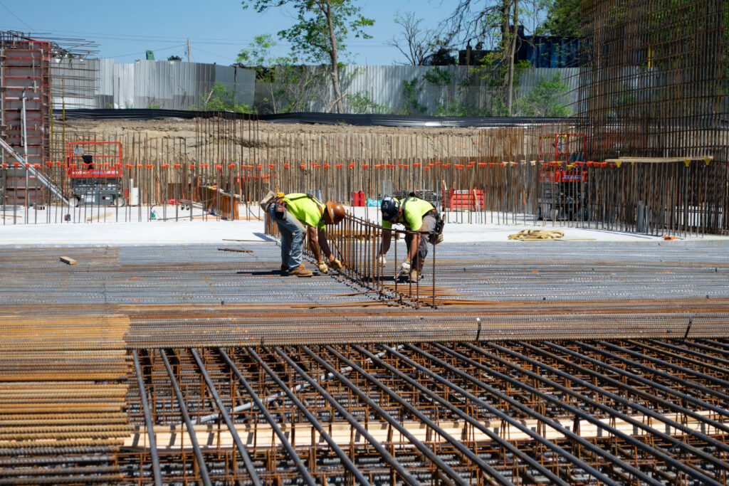 Two construction workers wearing safety gear bend over to secure steel rebar on a large concrete foundation at a building site, surrounded by metal rods and construction equipment.