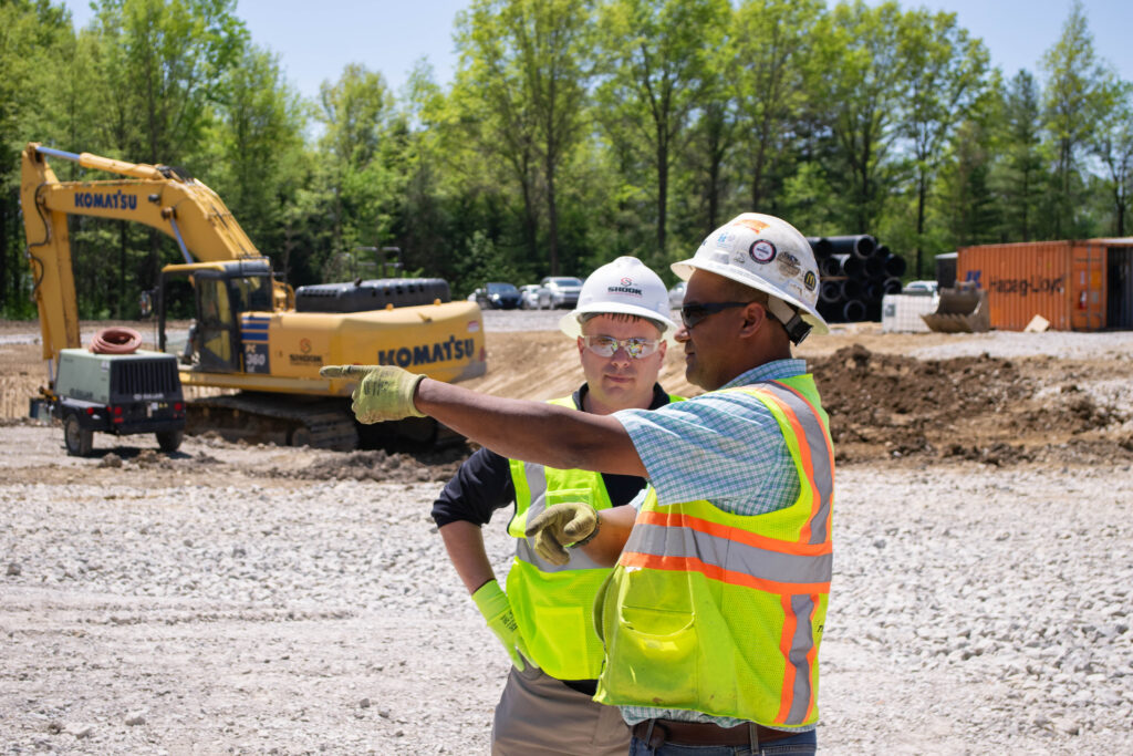 Two construction workers in safety vests and helmets talk at a construction site with equipment, gravel, and trees in the background. One worker points while the other listens.