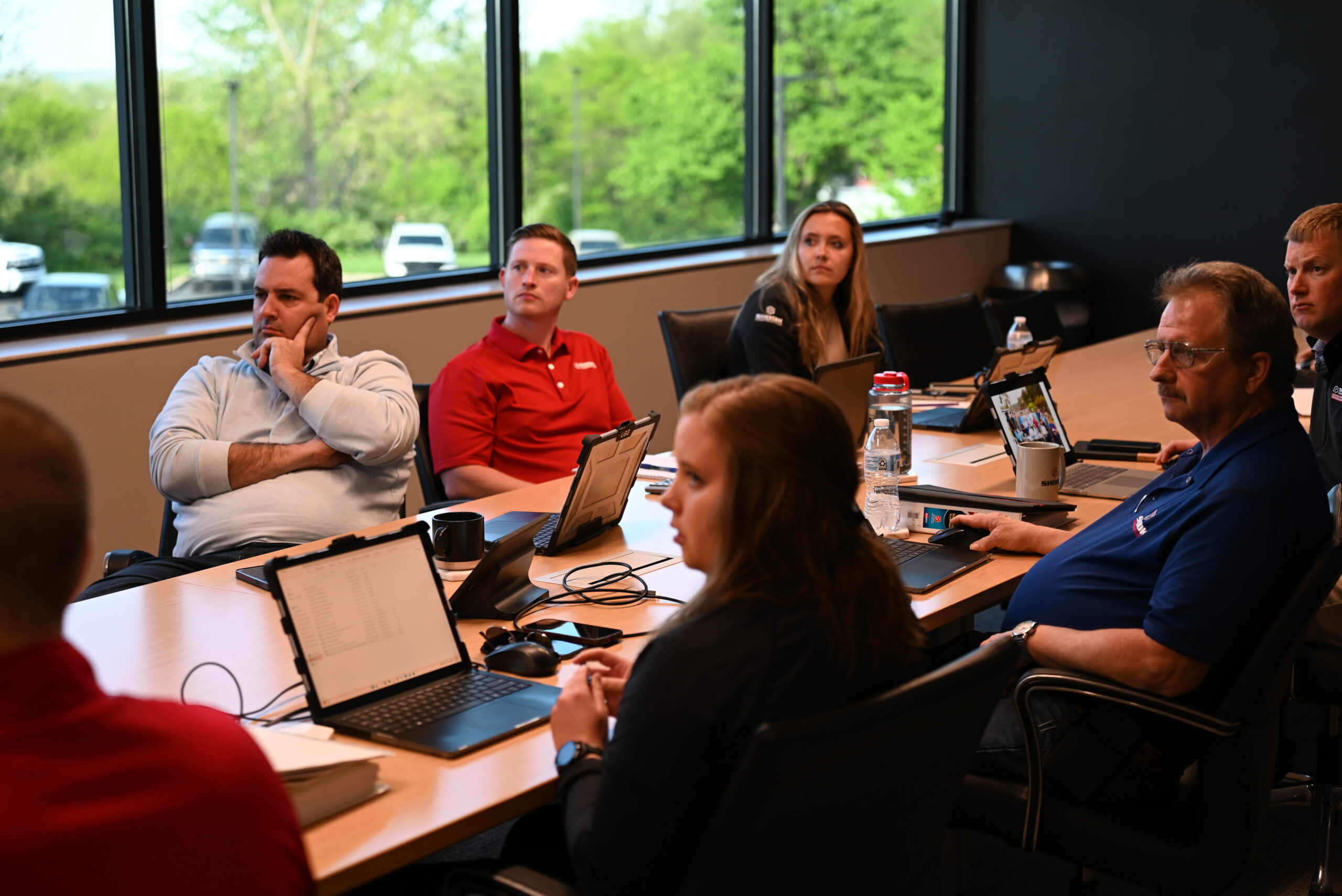 Six people sit around a conference table with laptops and tablets, attentively listening during a meeting in a bright room with large windows showing greenery outside.