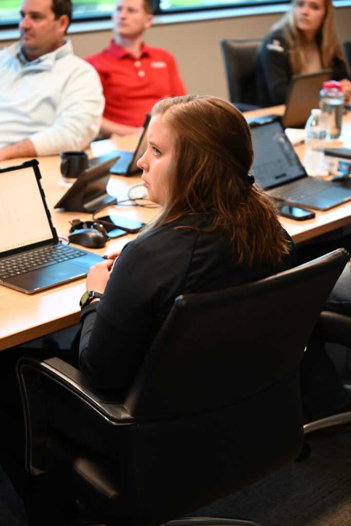 Woman with brown hair sits at a conference table with a laptop, listening attentively. Others are seated around her, focused on a meeting.
