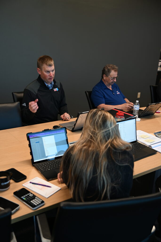Three people sit at a conference table with laptops and papers. One man in black speaks while another man in blue works on his laptop. A woman with long hair faces away, focused on her screen.
