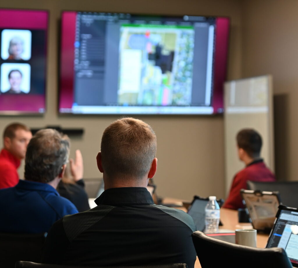 People seated around a conference table watch a large screen displaying a map or diagram. Laptops, coffee cups, and water bottles are on the table. The audience faces the screen attentively in a meeting room.