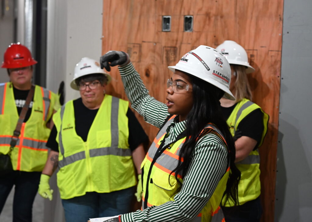 A woman in a hard hat and safety vest gestures while speaking to a group of construction workers wearing safety gear, standing indoors at a work site.