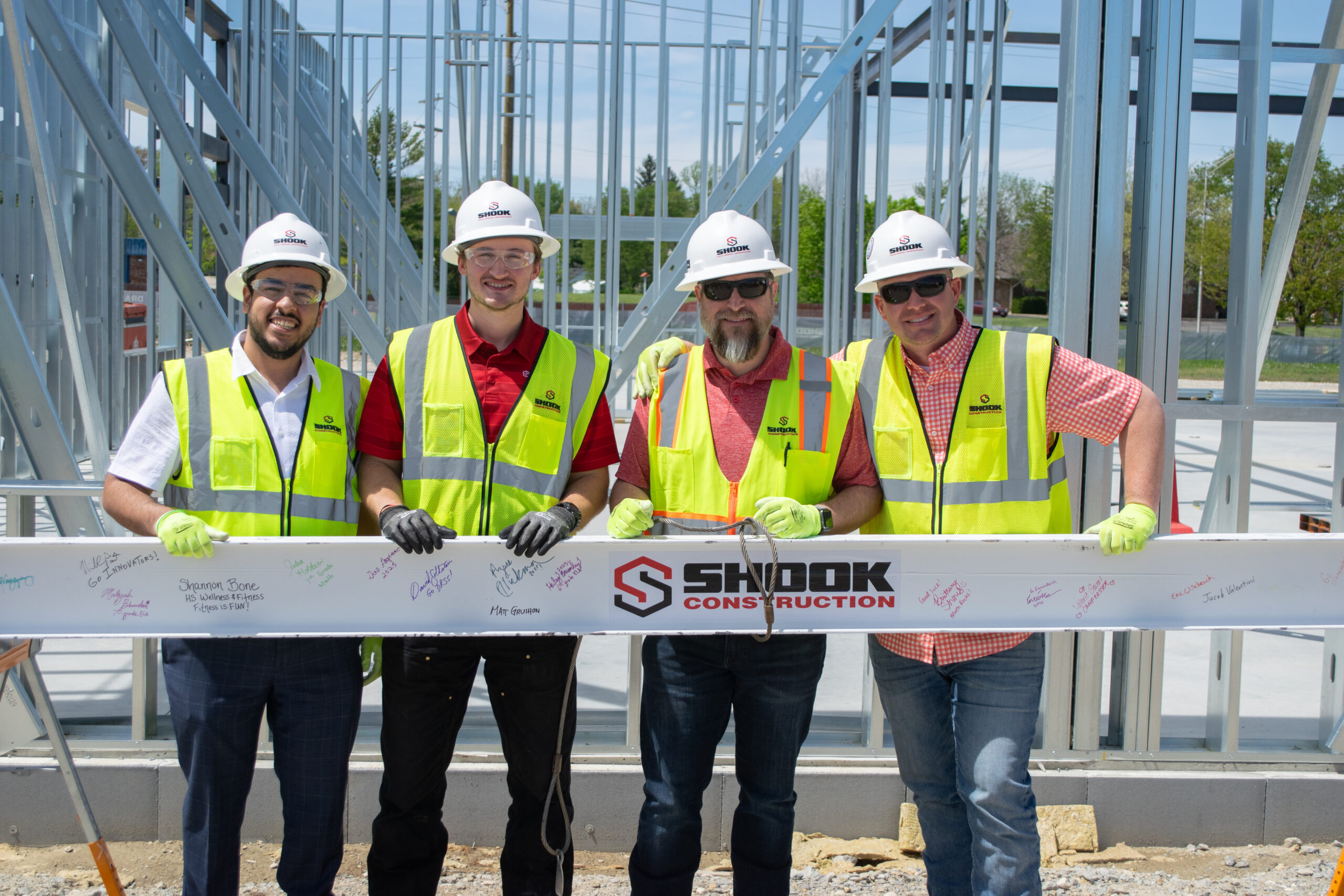Four men in safety vests and hard hats stand at a construction site, holding a signed beam with the Shook Construction logo. The buildings metal framework is visible in the background.