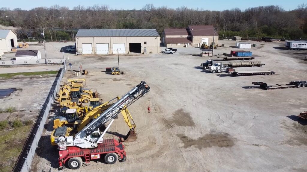 Construction yard with several yellow vehicles, a large red and white crane, and equipment parked on dirt ground. There are warehouse buildings, trucks, and trees in the background under a clear sky.