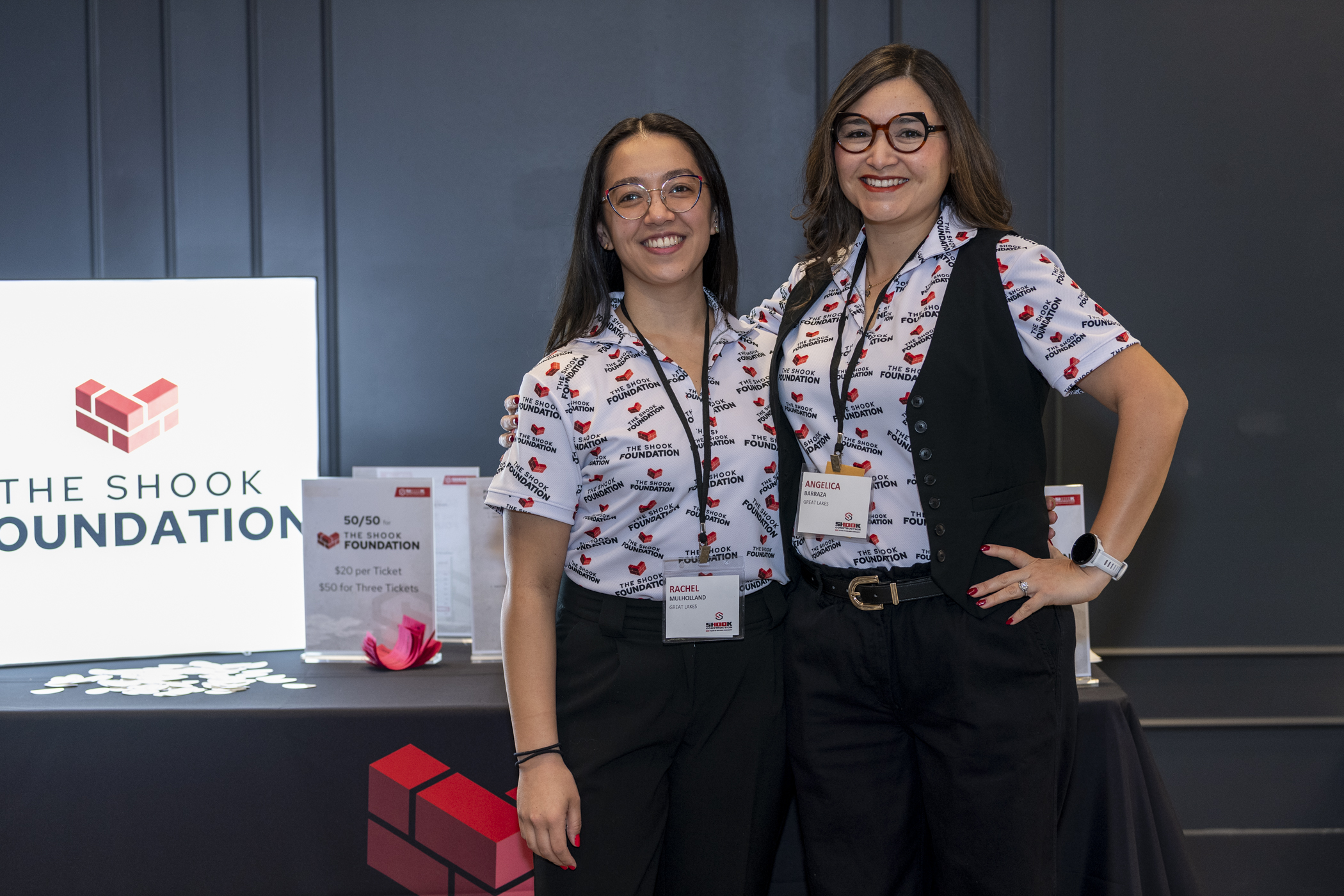 Two women wearing matching logo shirts stand smiling at a booth for The Shook Foundation. One has a badge reading MICHEL, and the other ANGELA. A white sign with the foundations name and red logo is visible beside them.