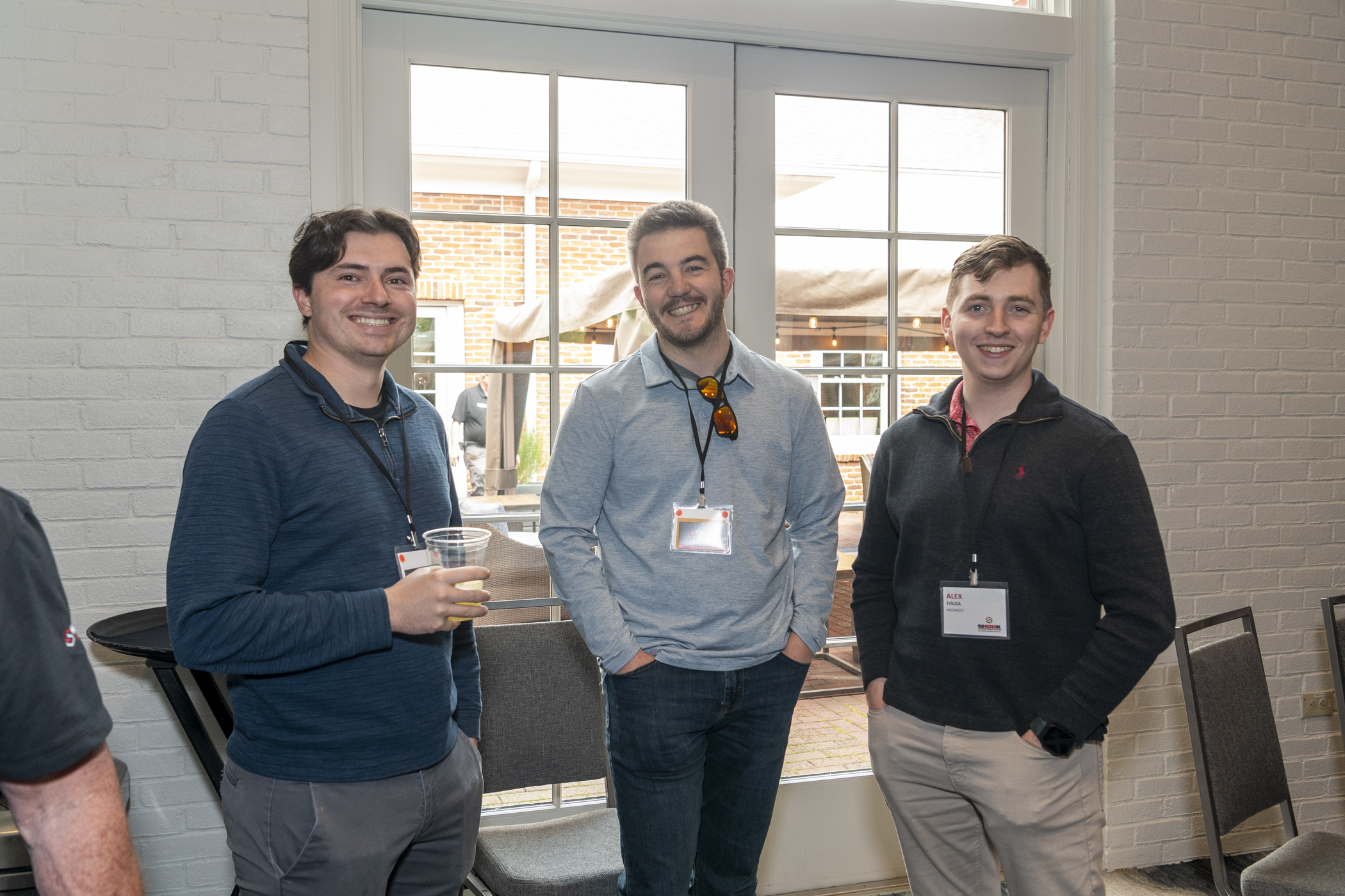 Three men stand indoors near a window, smiling at the camera. Two of them have drinks. All wear name badges and casual clothes. Outdoor scenery is visible through the glass doors behind them.