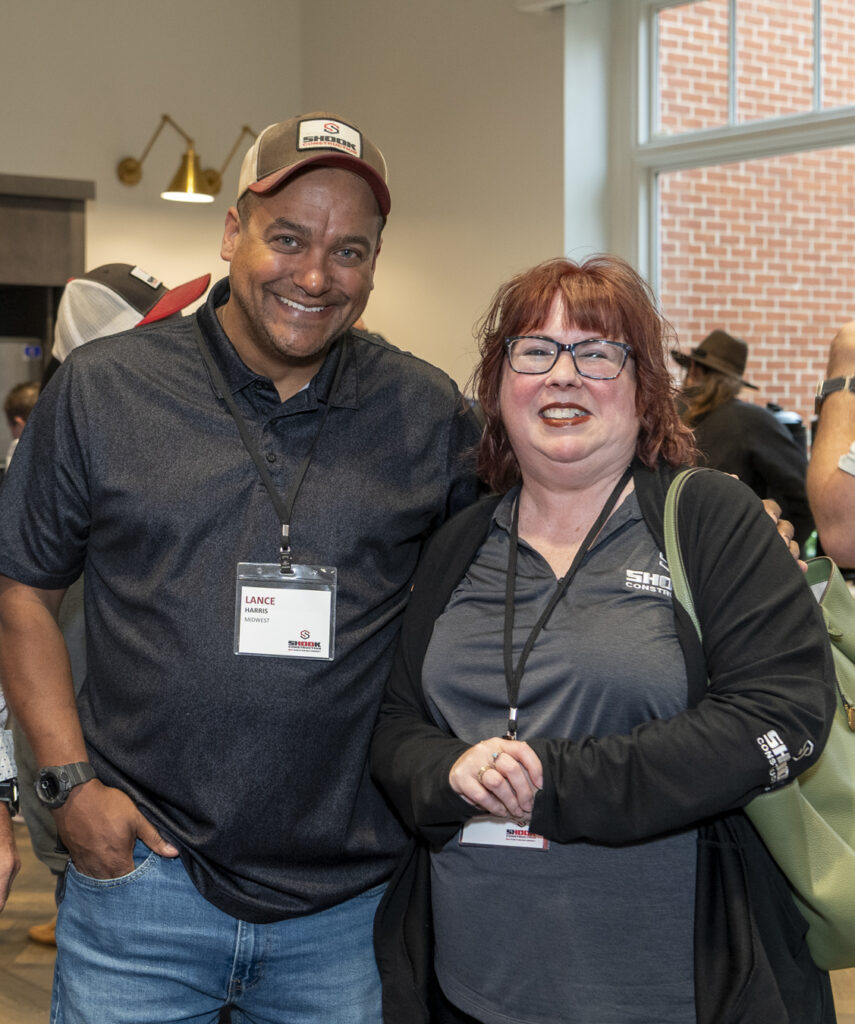 Three people wearing name tags stand close together indoors at a social event. They are smiling at the camera. Other people are mingling in the background. The setting appears casual and friendly.