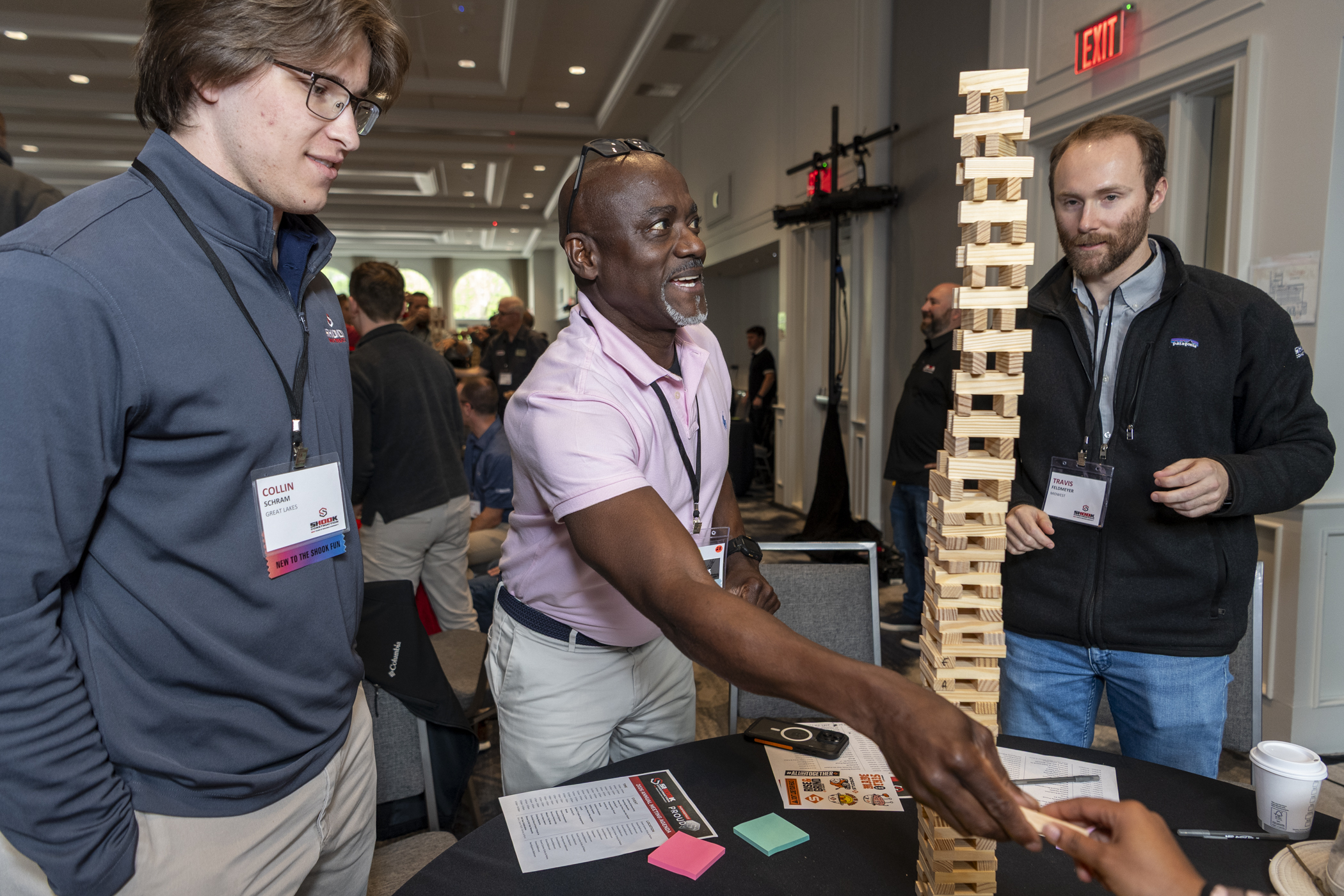 Three men stand around a tall Jenga tower at a table, one reaching to remove a block. The group appears focused and engaged, with others in the background in a conference room setting.
