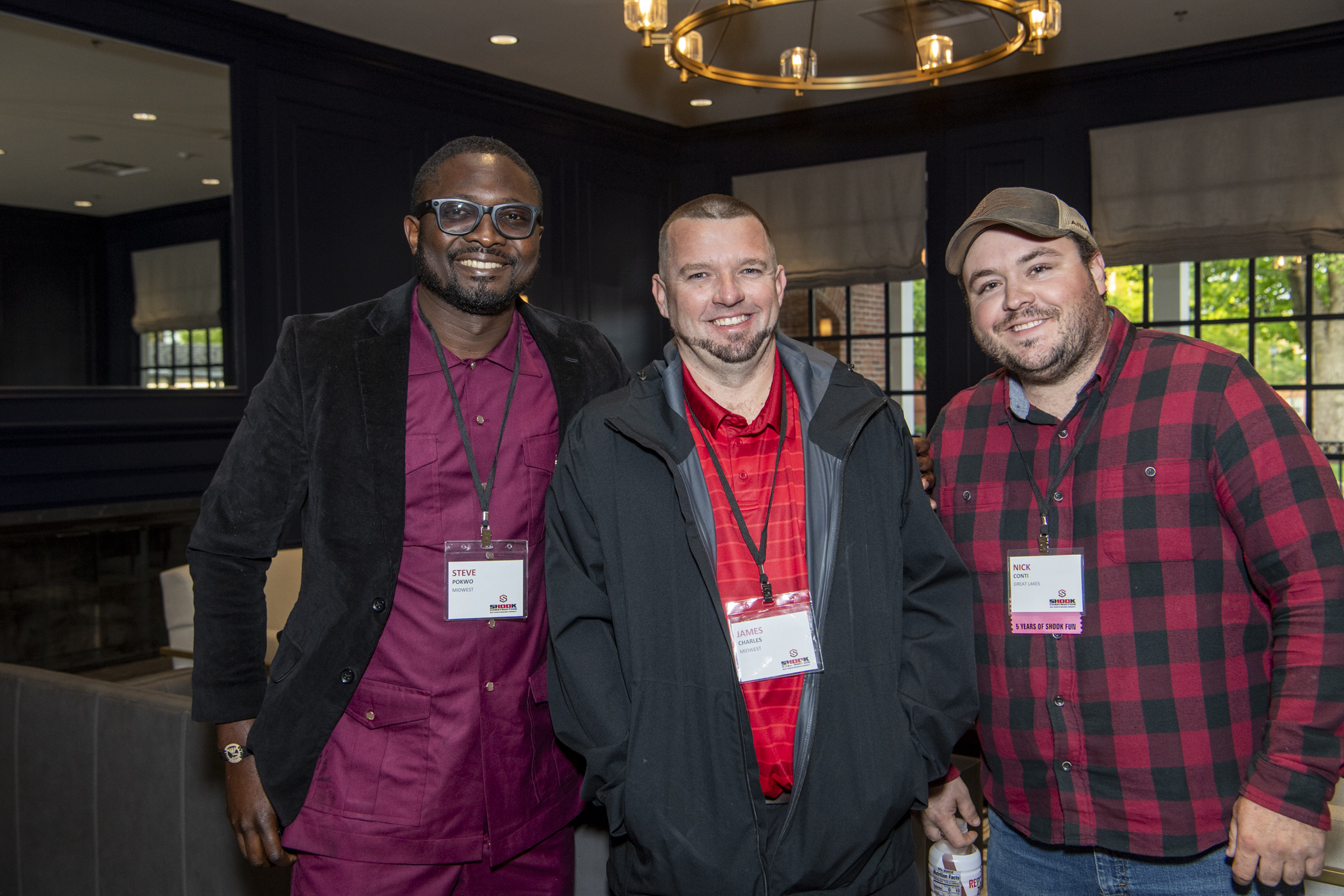 Three men stand together indoors at an event, all smiling for the camera and wearing name badges. They are dressed in business casual attire with red and black tones. A chandelier and windows are visible in the background.