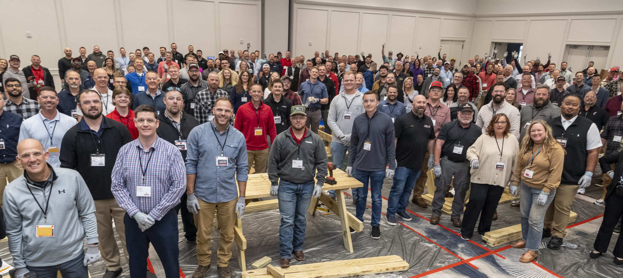 A large group of people, many wearing name tags, stand together in a conference room with wooden planks and assembly materials on tables, smiling at the camera.