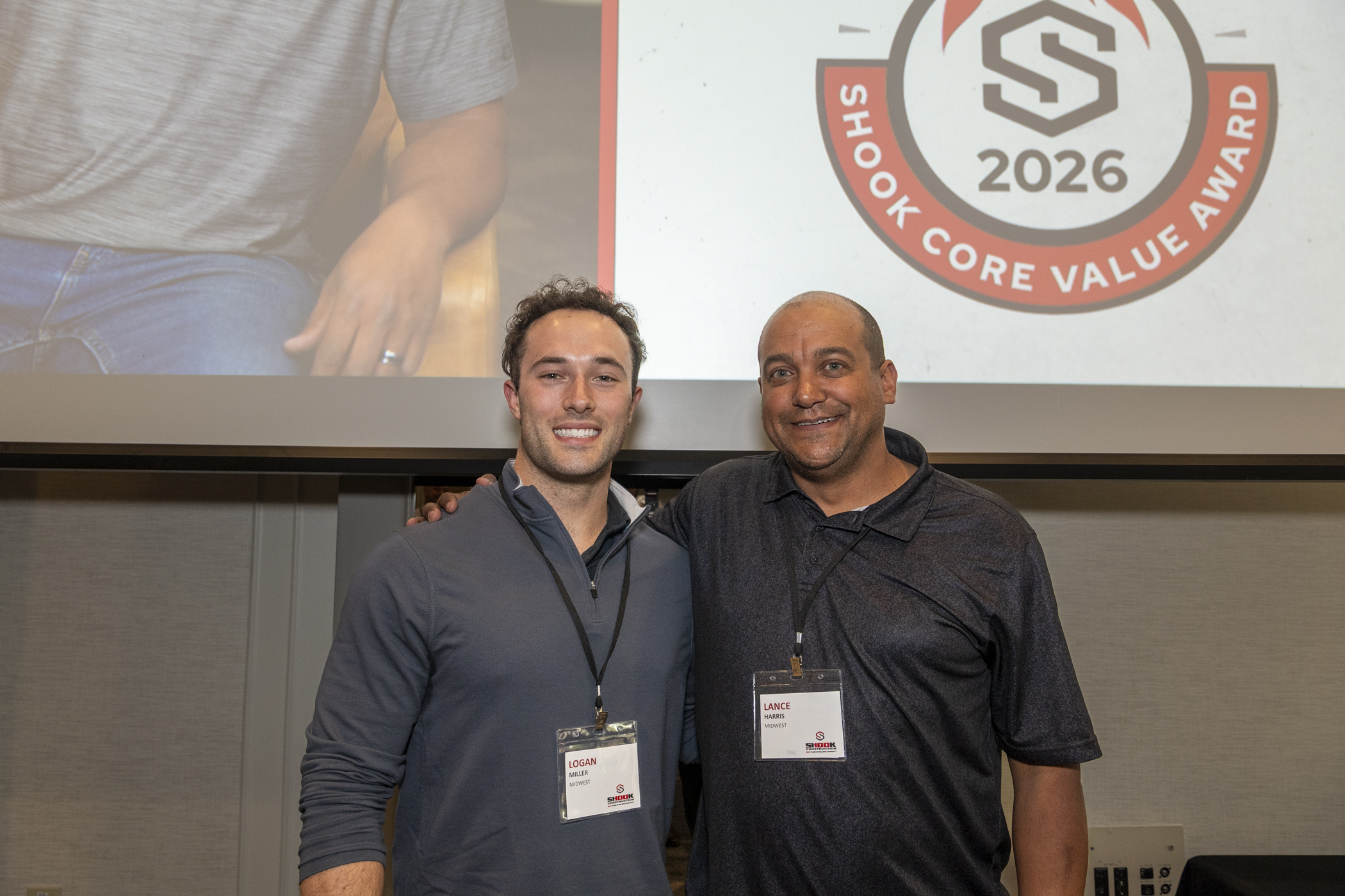 Two men smiling and posing together at an event; both wear name badges. Behind them is a screen featuring the Shook Core Value Award 2026 logo.