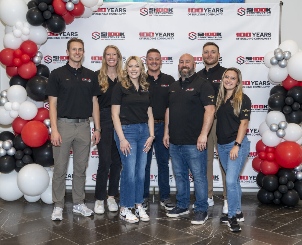 Seven people stand smiling in front of a Shook Construction 100 Years banner, surrounded by red, white, black, and silver balloons. All wear black company shirts and casual pants, standing on a shiny tiled floor.