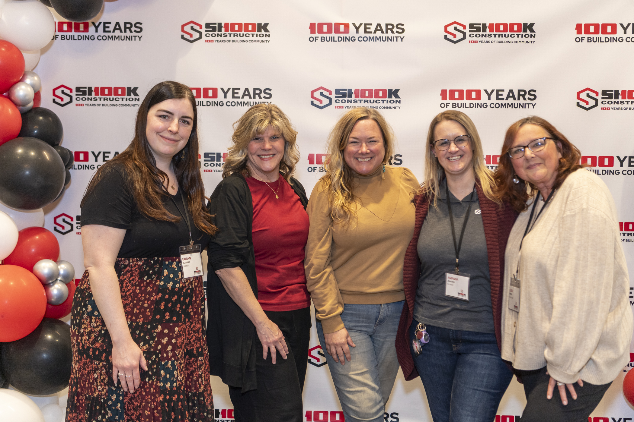 Five women stand smiling in front of a step-and-repeat banner for Shook Constructions 100th anniversary, with black, white, and red balloons to the left.