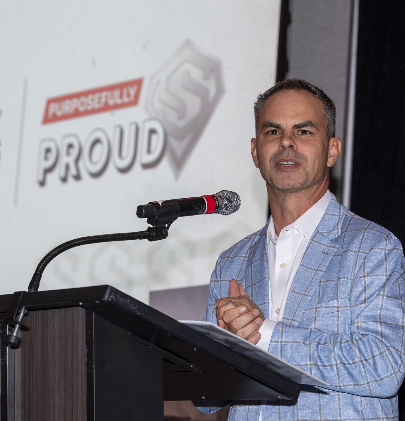 A man in a light blue plaid suit speaks at a podium with a microphone. Behind him, a screen displays the words Purposefully Proud with a stylized S logo.