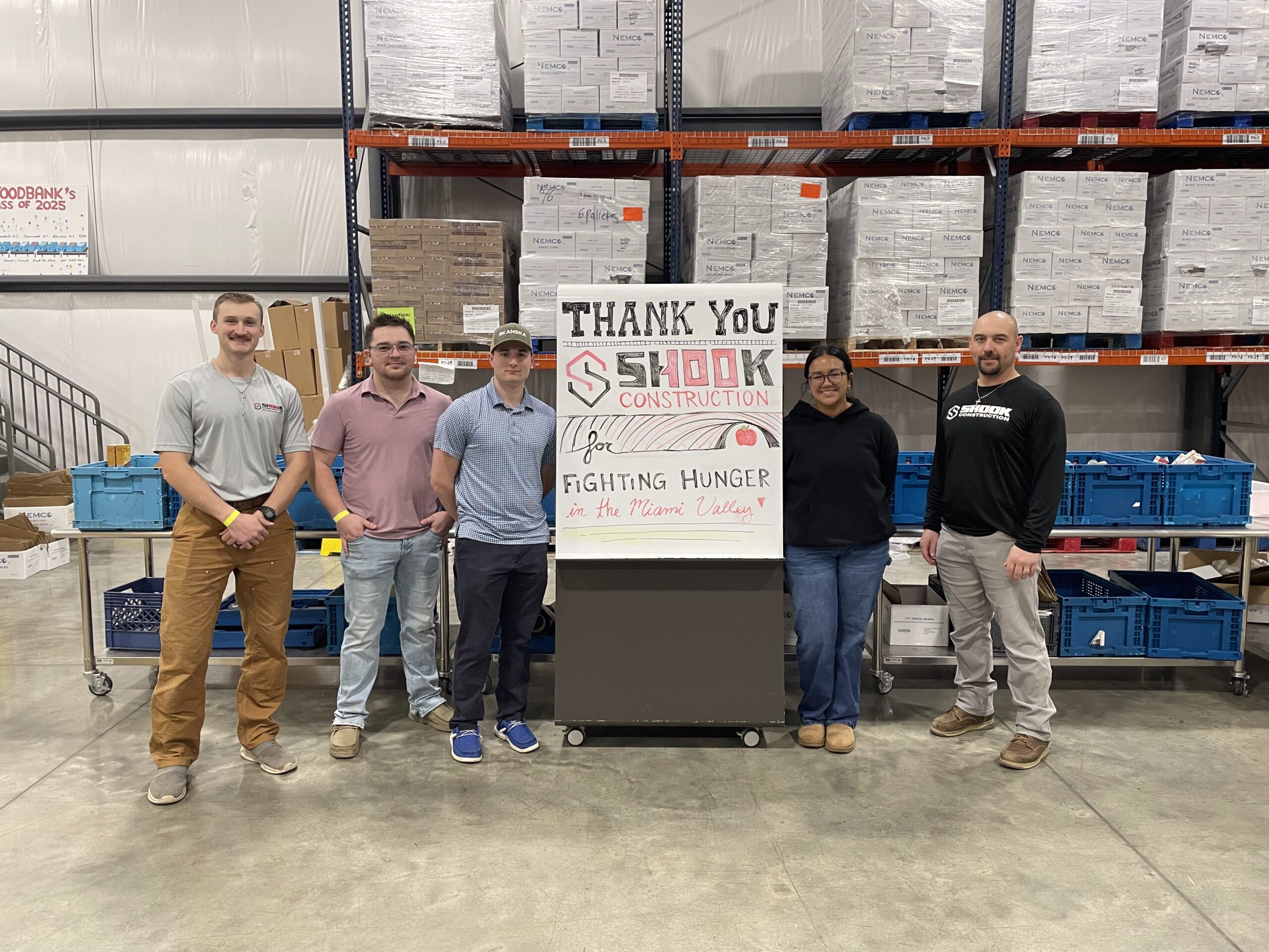 Six people stand in a warehouse behind a sign thanking Shook Construction for fighting hunger. There are boxes and blue bins on shelves and tables around them.