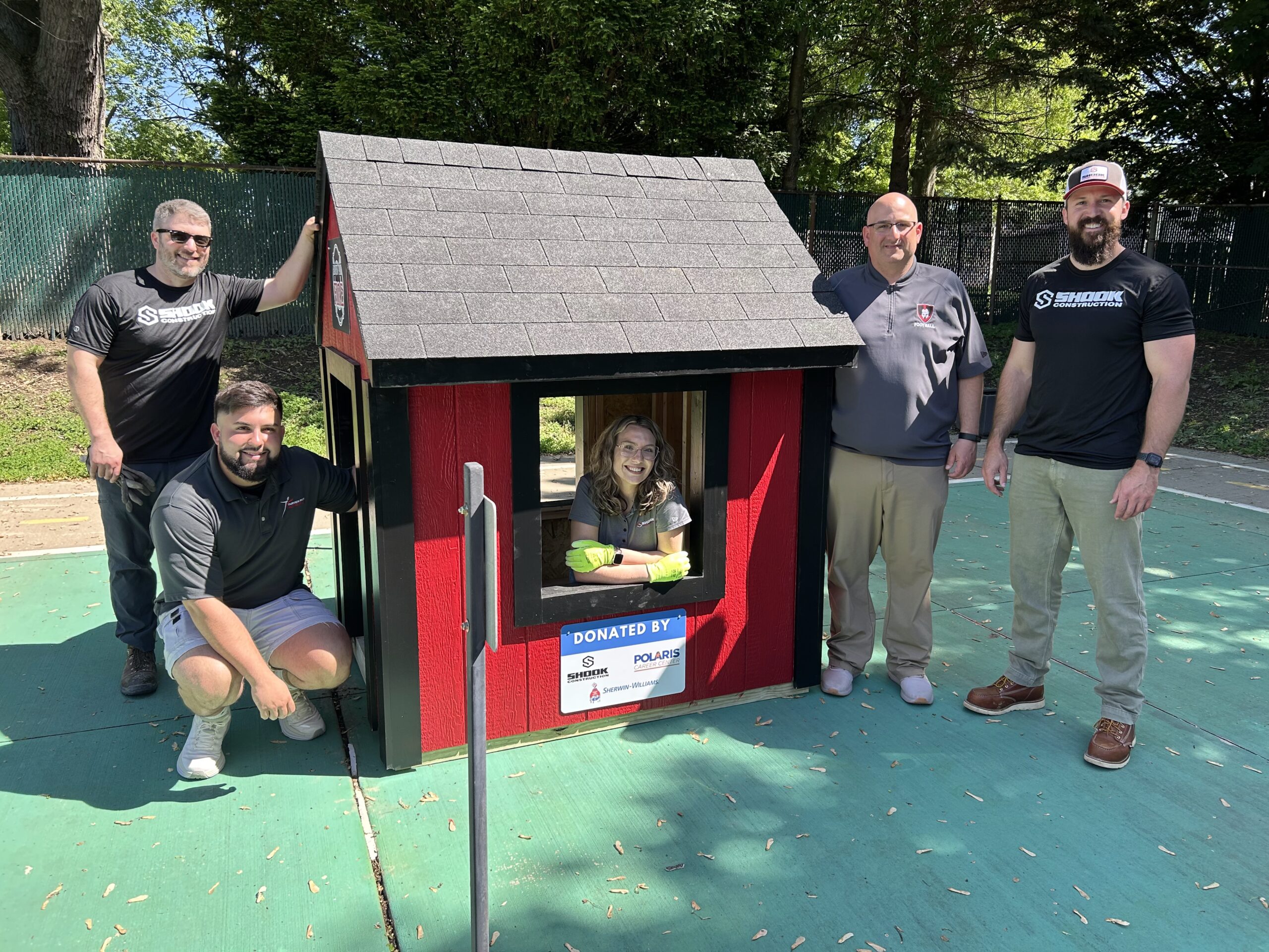 Five adults stand around a small red playhouse on an outdoor court; one person sits inside smiling. A donation sign is displayed on the playhouse, and trees are visible in the background.