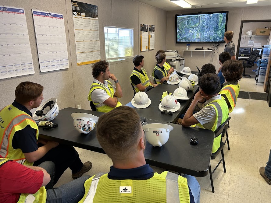 A group of workers in safety vests sit at tables with helmets, watching a presentation with a map displayed on a wall screen in an office setting.