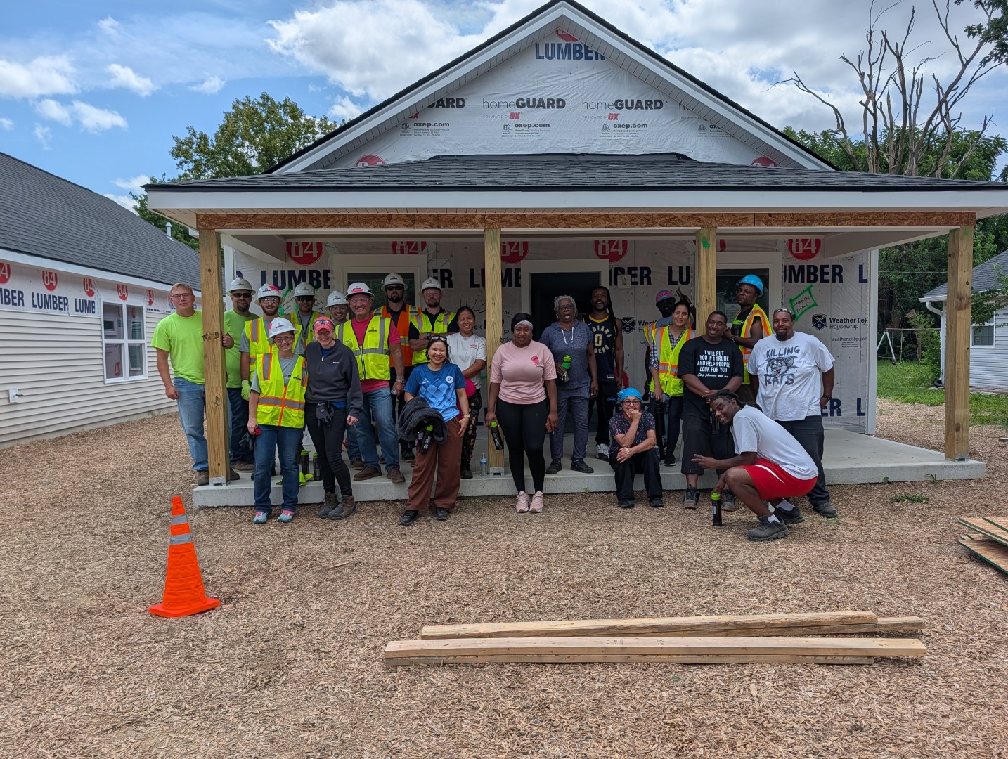 A diverse group of people, some in safety vests and hard hats, pose in front of a partially built house. Wooden planks and an orange cone are in the foreground. The sky is partly cloudy.