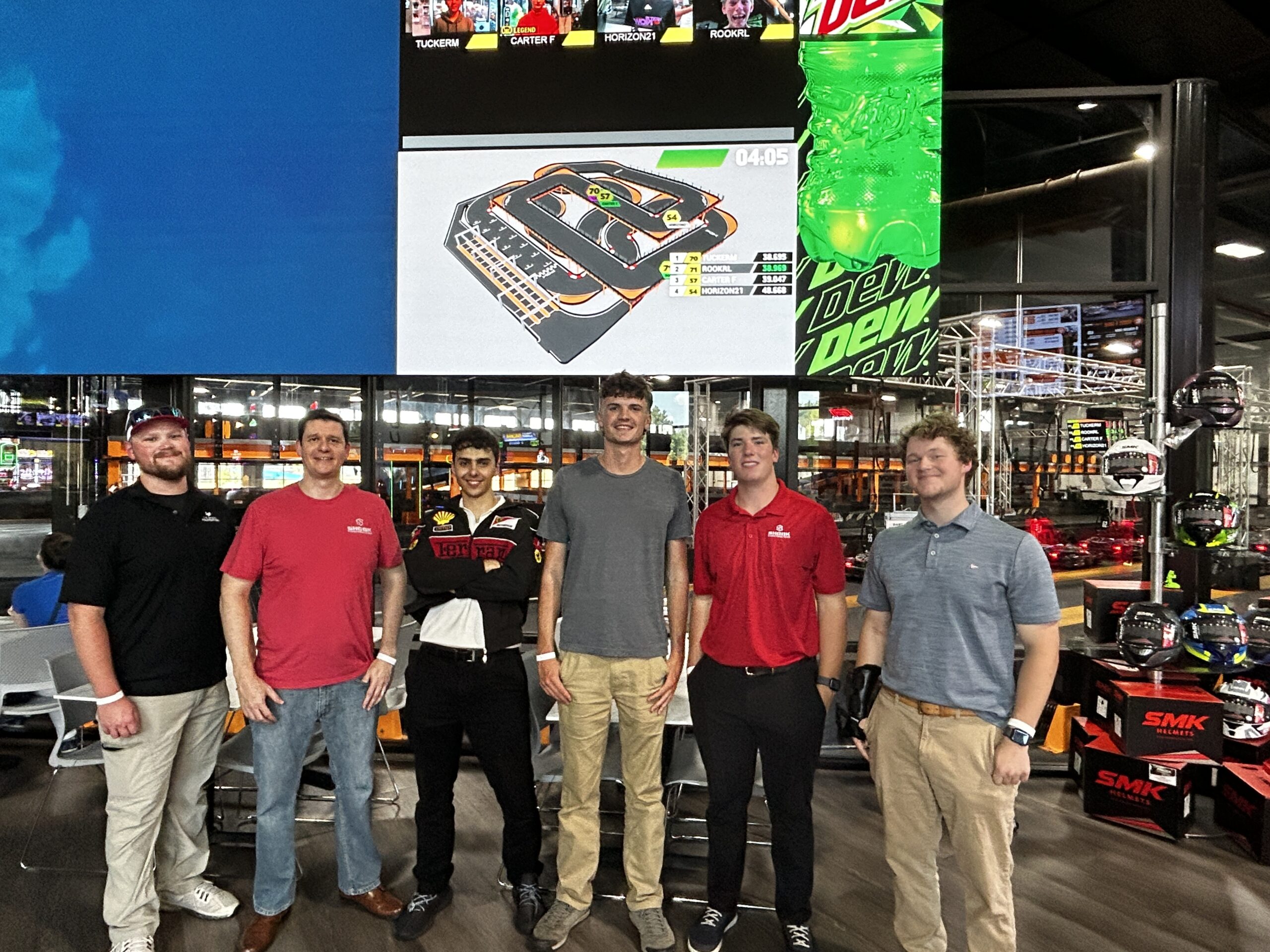 Six men stand in front of a large race track screen at an indoor go-kart facility, posing and smiling for the camera. Some wear casual outfits, while others wear racing suits or branded shirts. Racing helmets are displayed nearby.