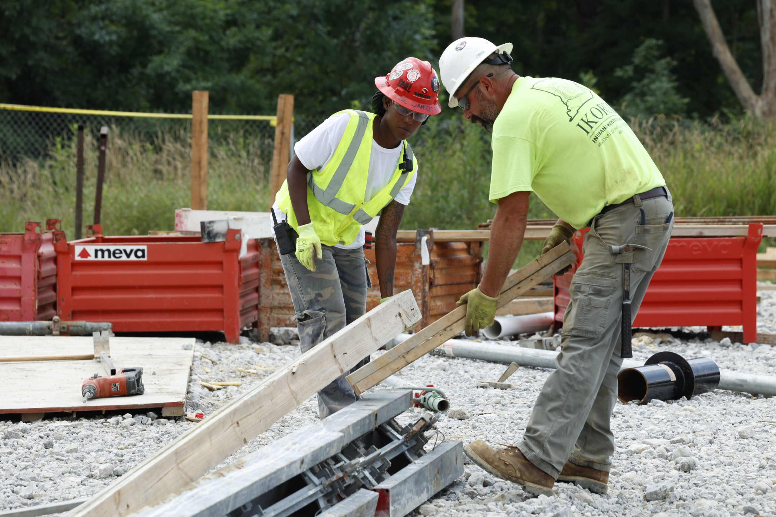 Two construction workers in safety gear lift wooden planks at a building site with gravel on the ground and red barriers in the background.