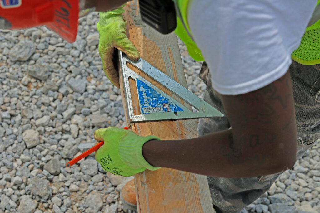 A construction worker wearing gloves uses a speed square and pencil to mark a piece of wood on a gravel-covered site.