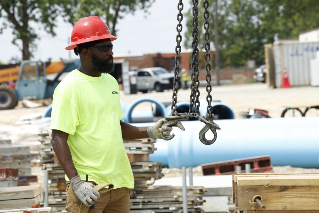 A construction worker in a neon shirt and red hard hat guides a large metal hook on chains at a worksite, surrounded by equipment and materials.