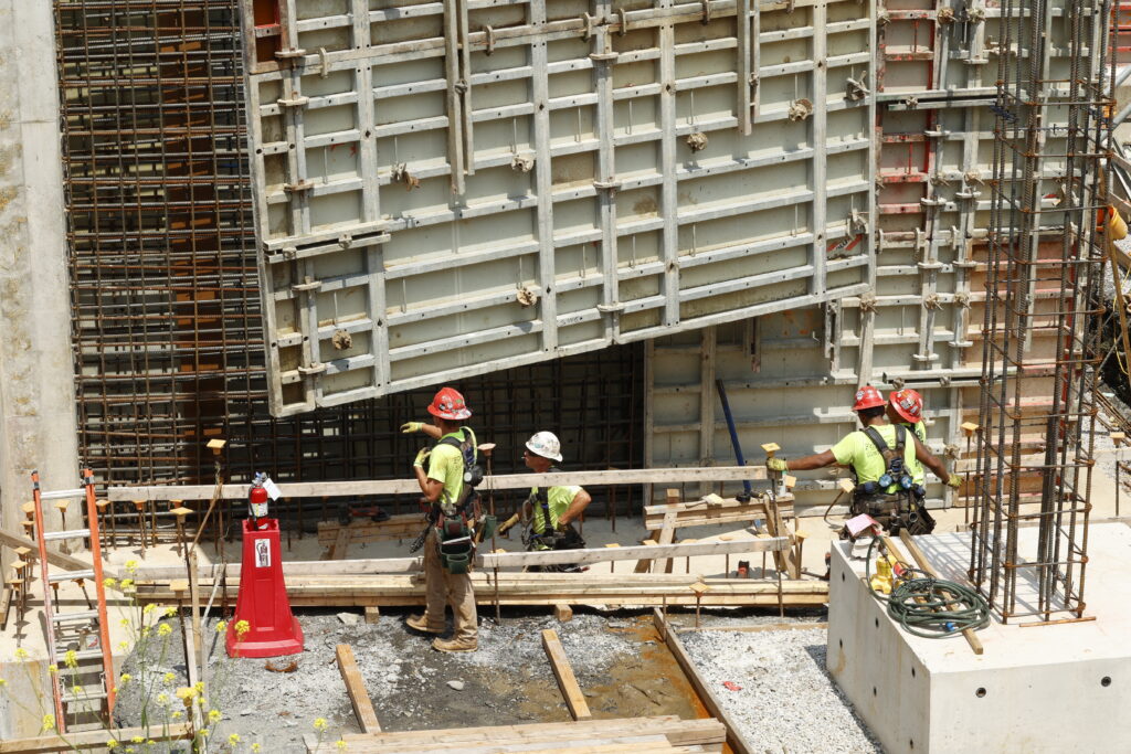 Three construction workers in safety gear guide a large metal panel into place on a building site surrounded by rebar, wood planks, and construction materials.