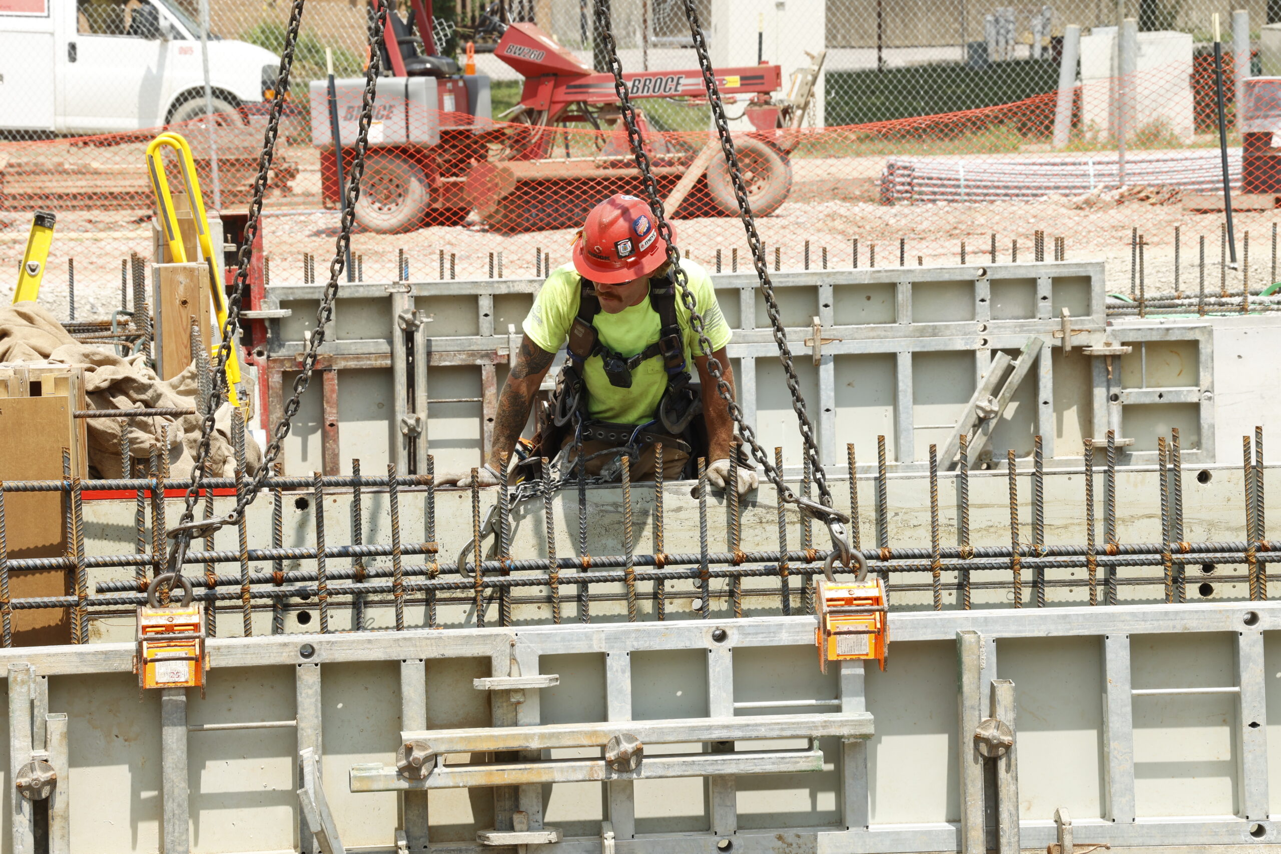 A construction worker in a red hard hat secures metal chains to a large concrete formwork, surrounded by steel rebar, at an outdoor building site. Construction equipment and materials are visible in the background.