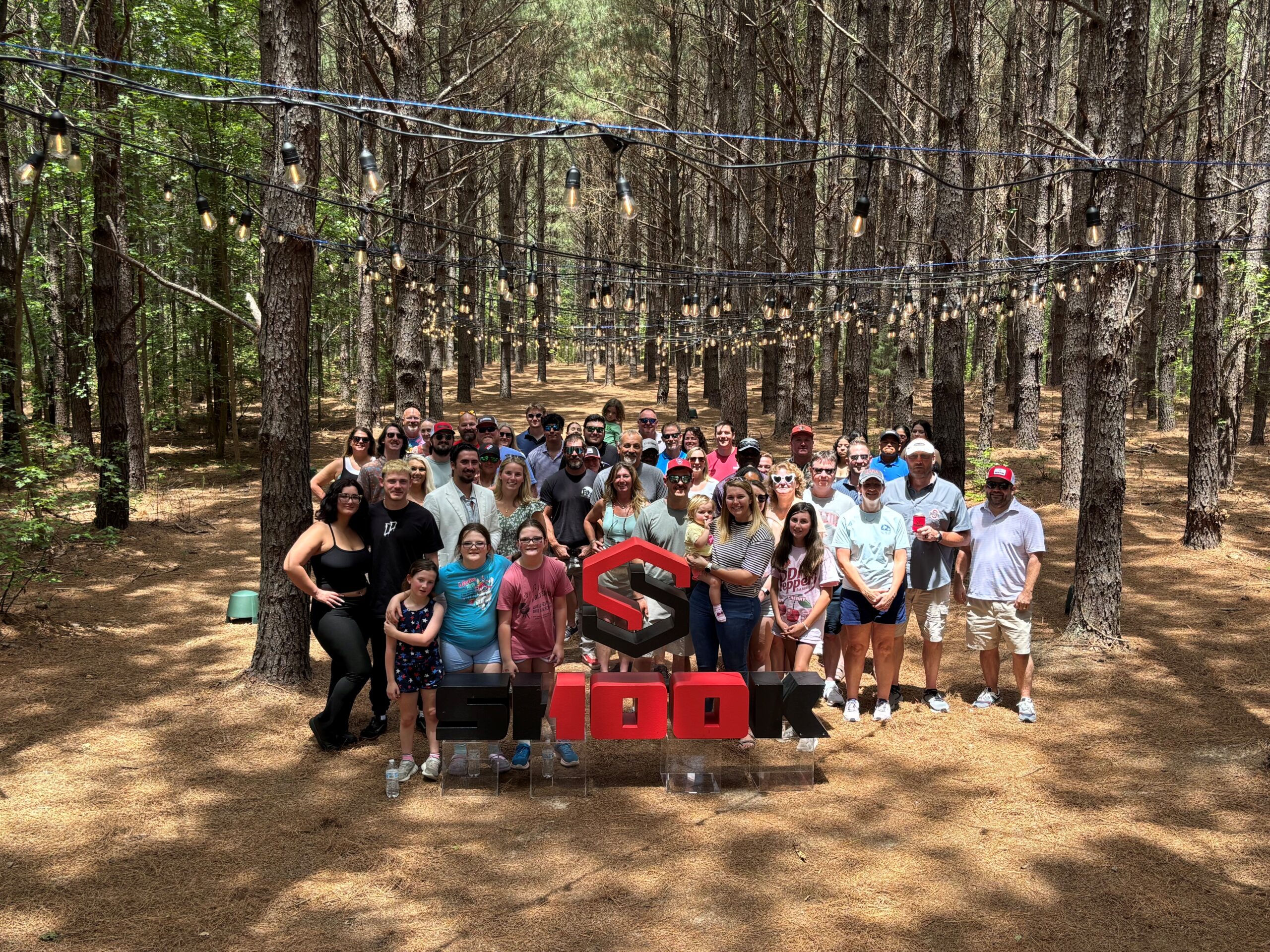 A group of people stands in a pine forest under string lights, posing behind a large red and black SHOOK sign. Sunlight filters through the trees, creating a festive, outdoor gathering atmosphere.