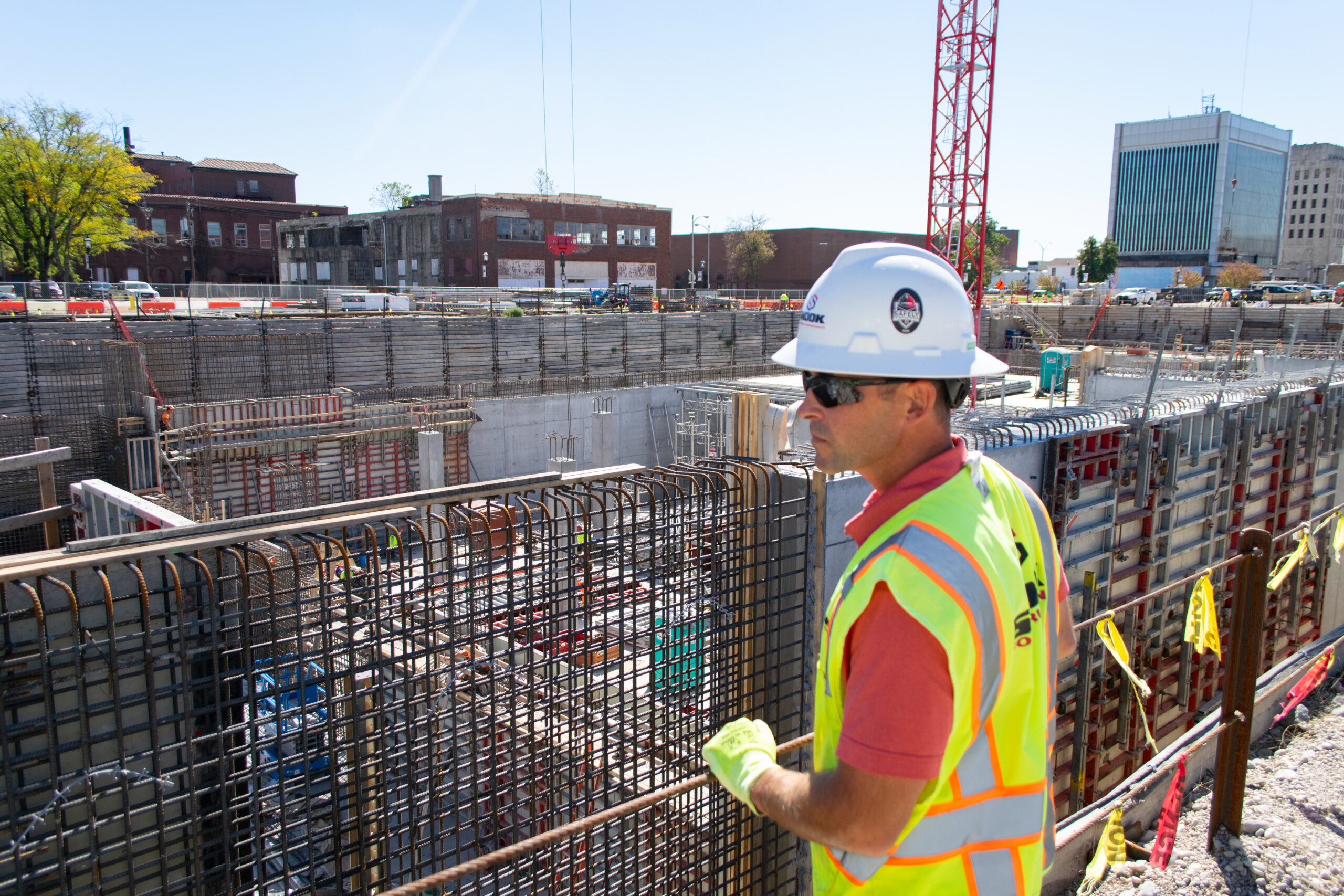 A construction worker in a hard hat and safety vest stands at a building site, observing reinforced steel bars and concrete forms under bright sunlight.