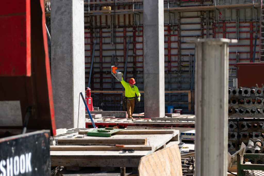 A construction worker in a yellow shirt and red helmet stands on a busy building site, holding up an orange item amid concrete columns and scattered materials.