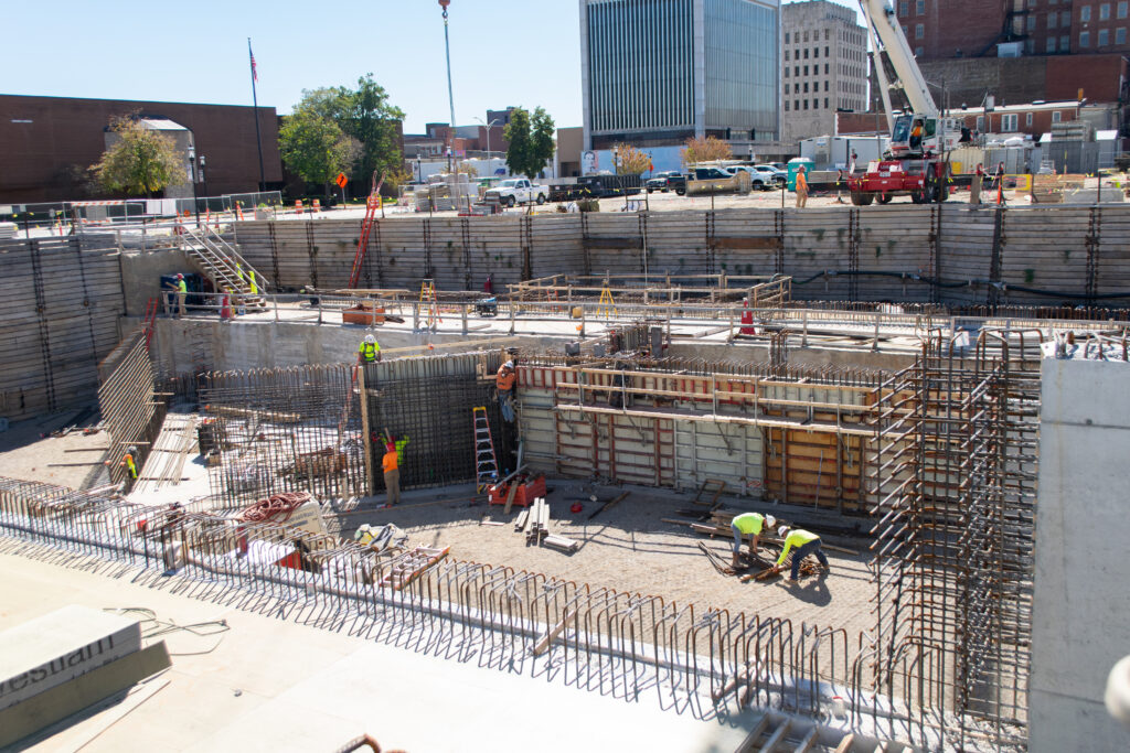 Construction site with workers, steel reinforcements, and heavy equipment within a deep excavation pit, surrounded by buildings and a crane in the background.