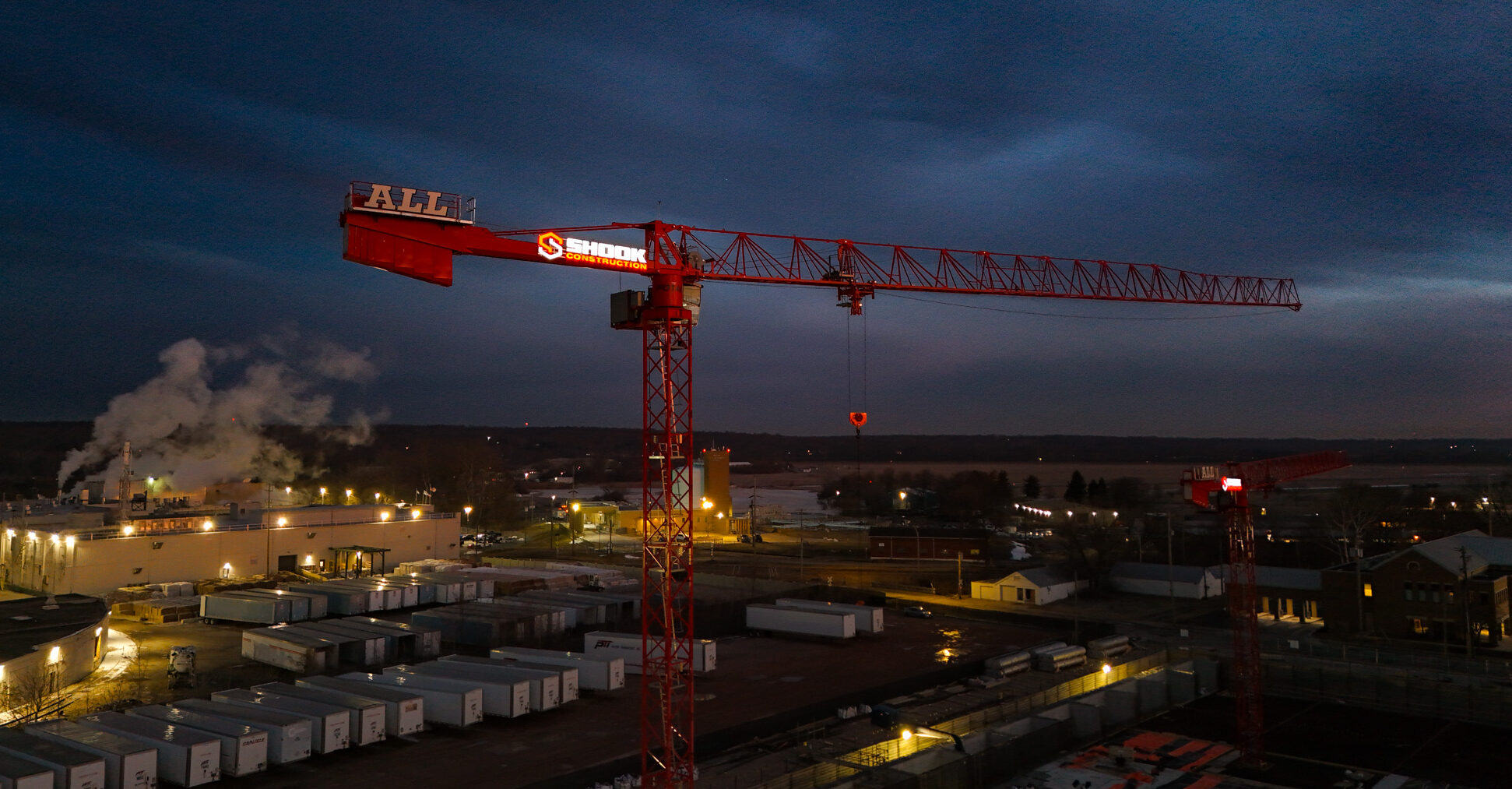 A large red construction crane stands over an industrial area at dusk, with factory buildings, steam rising, and trucks parked below. The sky is dark, and the crane is illuminated with red lights.
