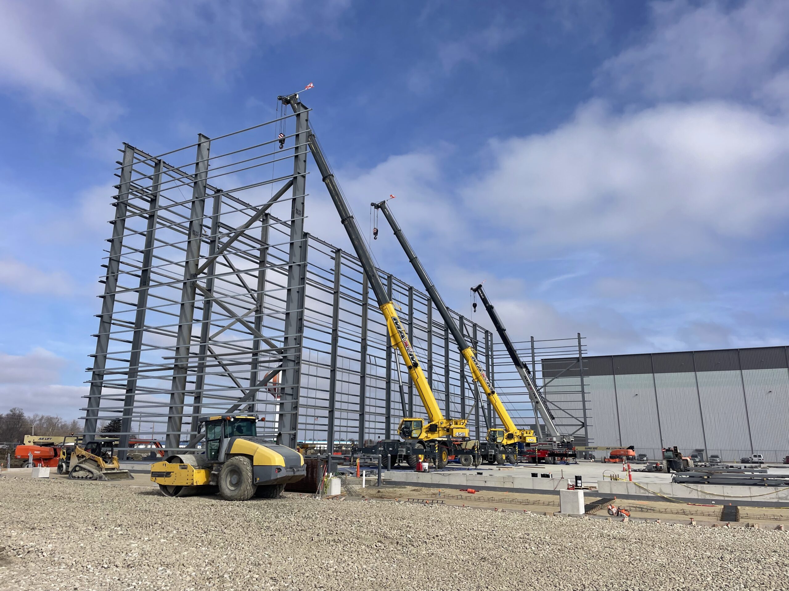 Construction site with large steel framework being assembled by two yellow cranes, adjacent to a finished gray building. Various construction vehicles and equipment are visible on gravel ground under a partly cloudy sky.